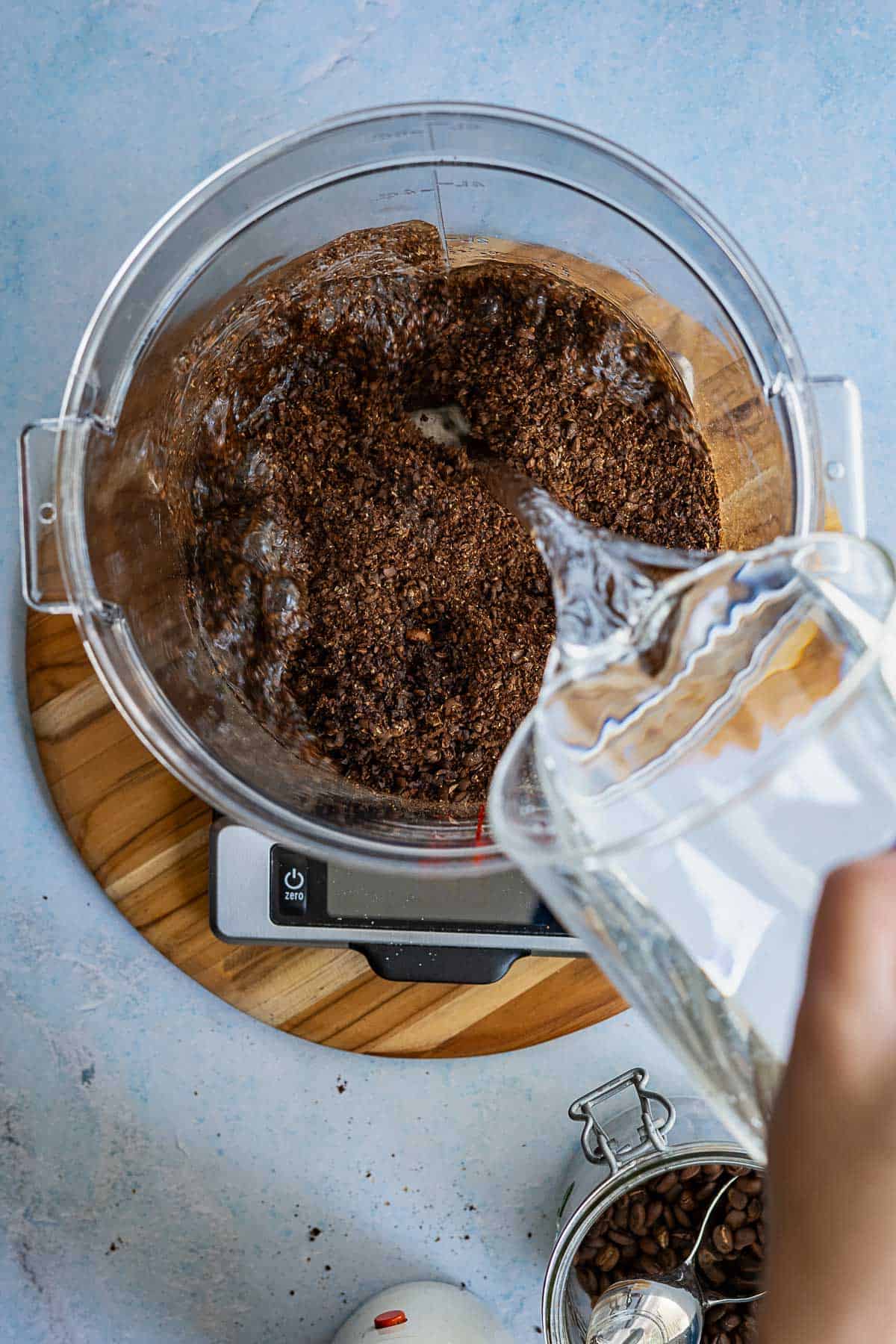 A hand pours water from a glass pitcher into a bowl of ground coffee, set on a wooden board and digital scale—a step in how to make cold brew. A jar of coffee beans and kitchen tools rest nearby on a light blue surface.