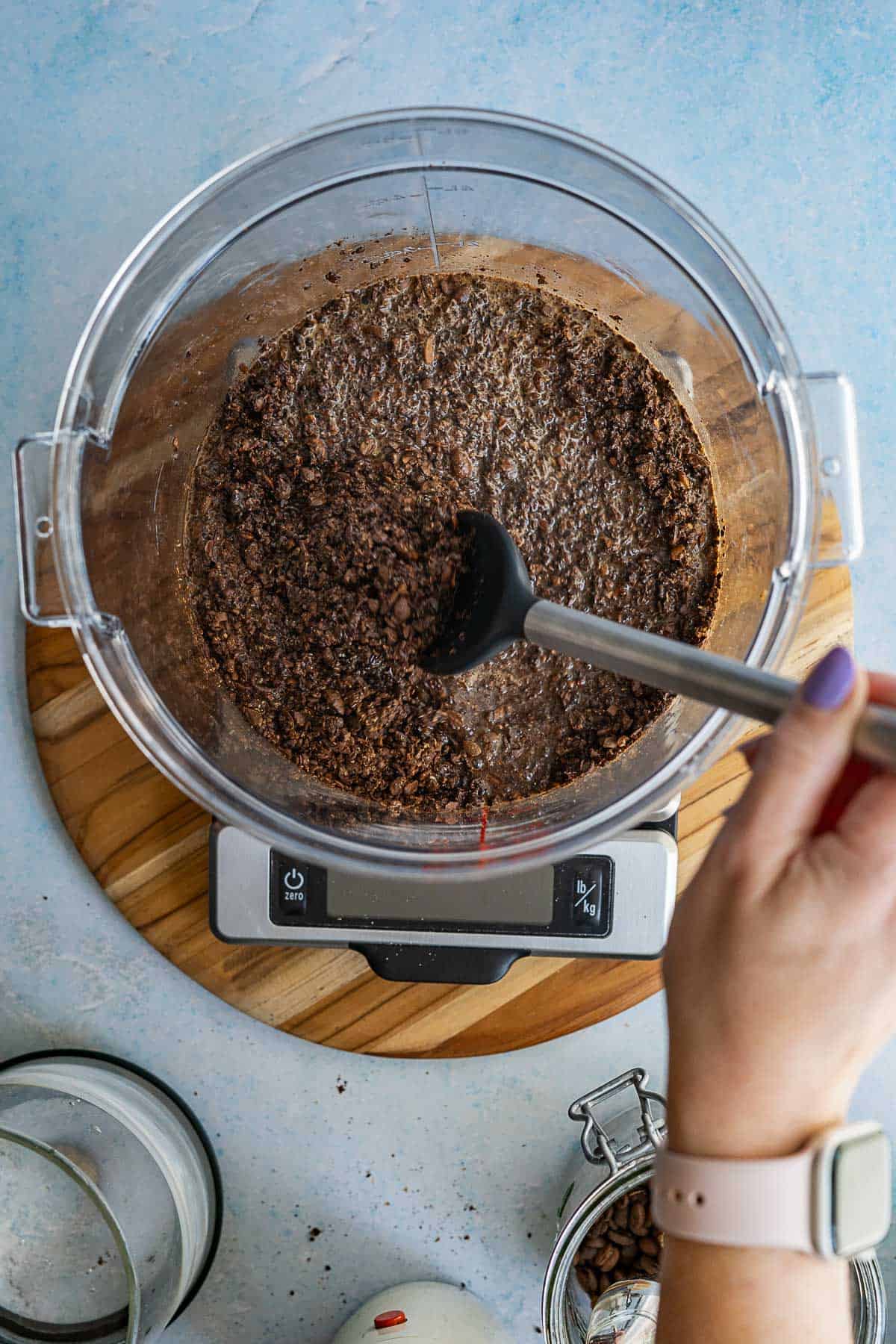 A hand stirs ground coffee and water in a food processor bowl, showing how to make cold brew. The bowl sits on a digital kitchen scale atop a wooden board, surrounded by kitchen items and coffee beans on a light blue surface.