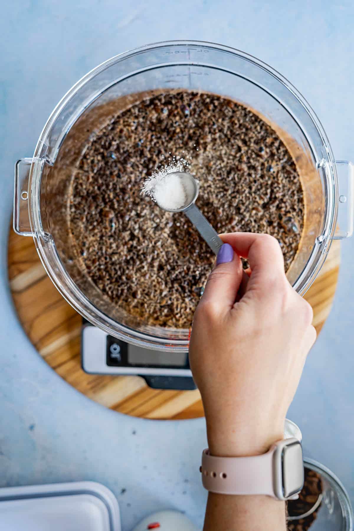 A person wearing a light pink watch holds a teaspoon of salt over a large clear bowl filled with a brown, grainy mixture, preparing to add it—perfect for showing how to make cold brew at home. The bowl sits on a wooden board.