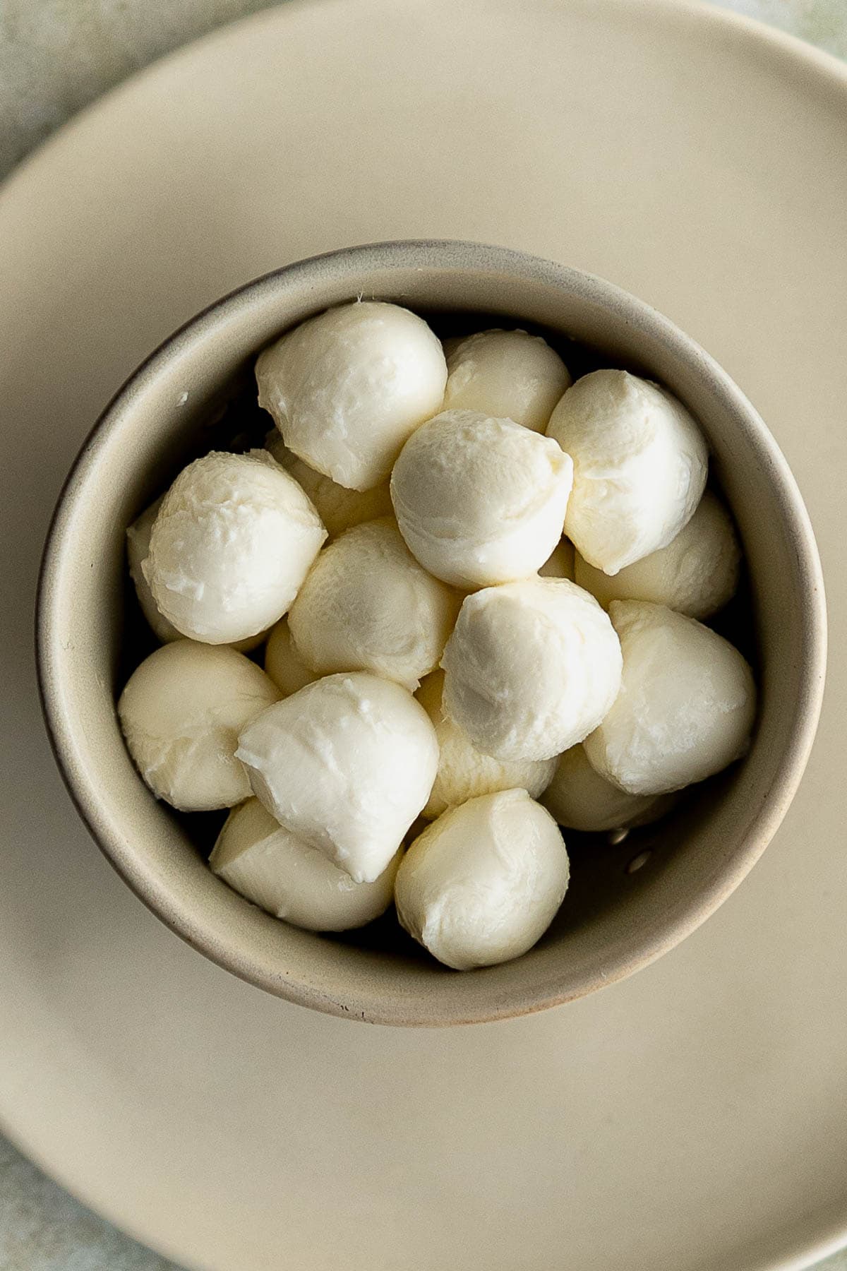 A bowl filled with small, round drained mozzarella cheese balls sits on a beige plate viewed from above, perfect for adding to a fresh mozzarella cheese and tomato salad.