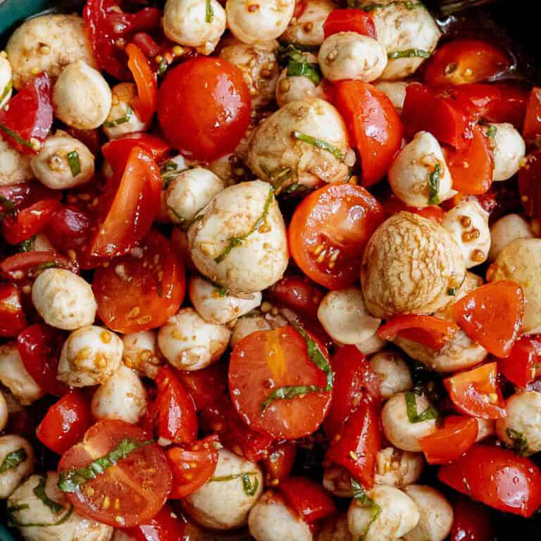 A close-up of a mozzarella cheese and tomato salad with cherry tomatoes, mozzarella balls, chopped basil, and balsamic dressing. The ingredients are mixed together, showcasing vibrant red, white, and green colors.