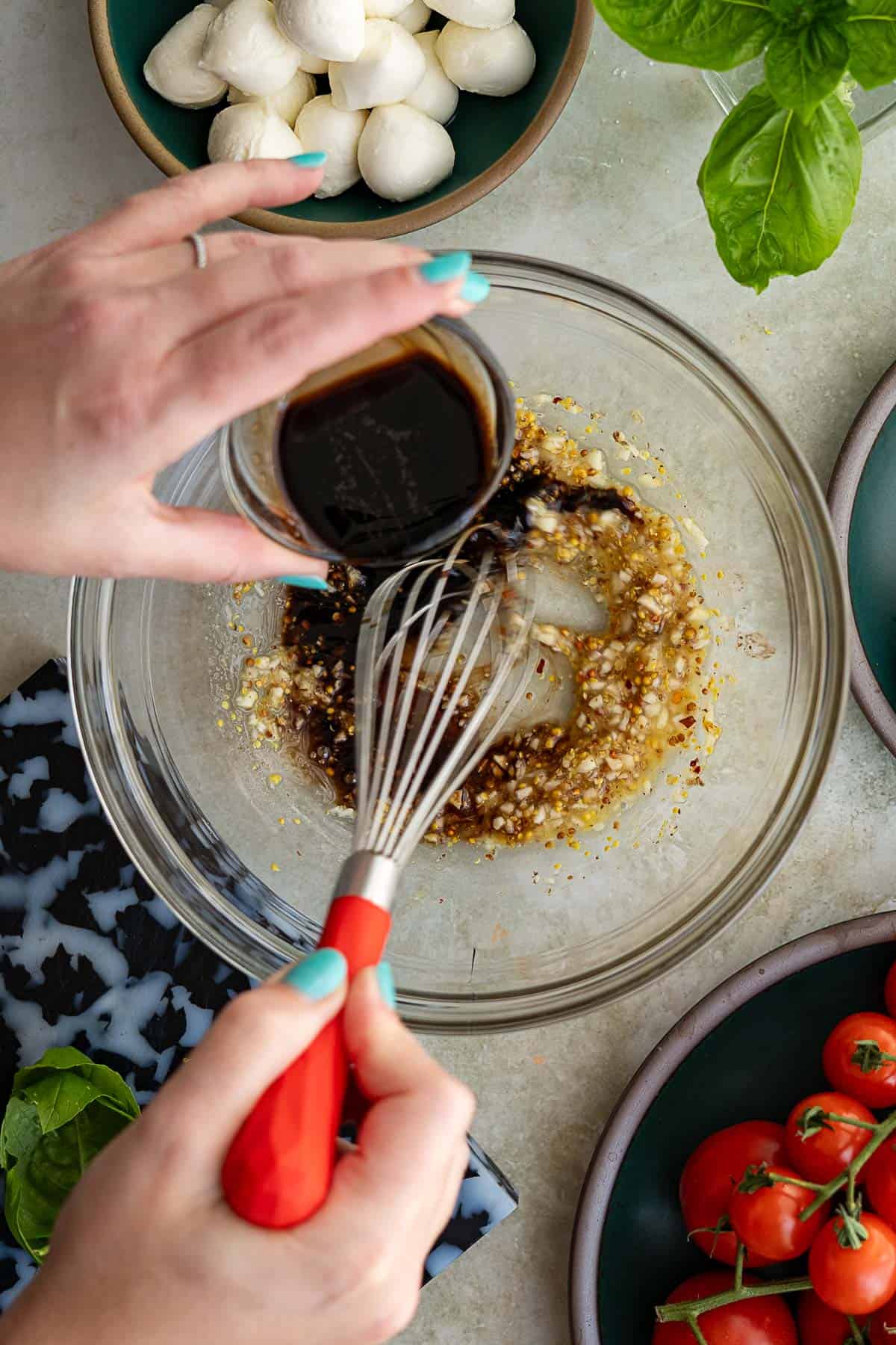 A person pours balsamic vinegar into a glass bowl of minced garlic and whisks with a red-handled whisk, preparing a dressing for mozzarella cheese and tomato salad. Fresh mozzarella balls, basil leaves, and tomatoes sit on plates nearby.