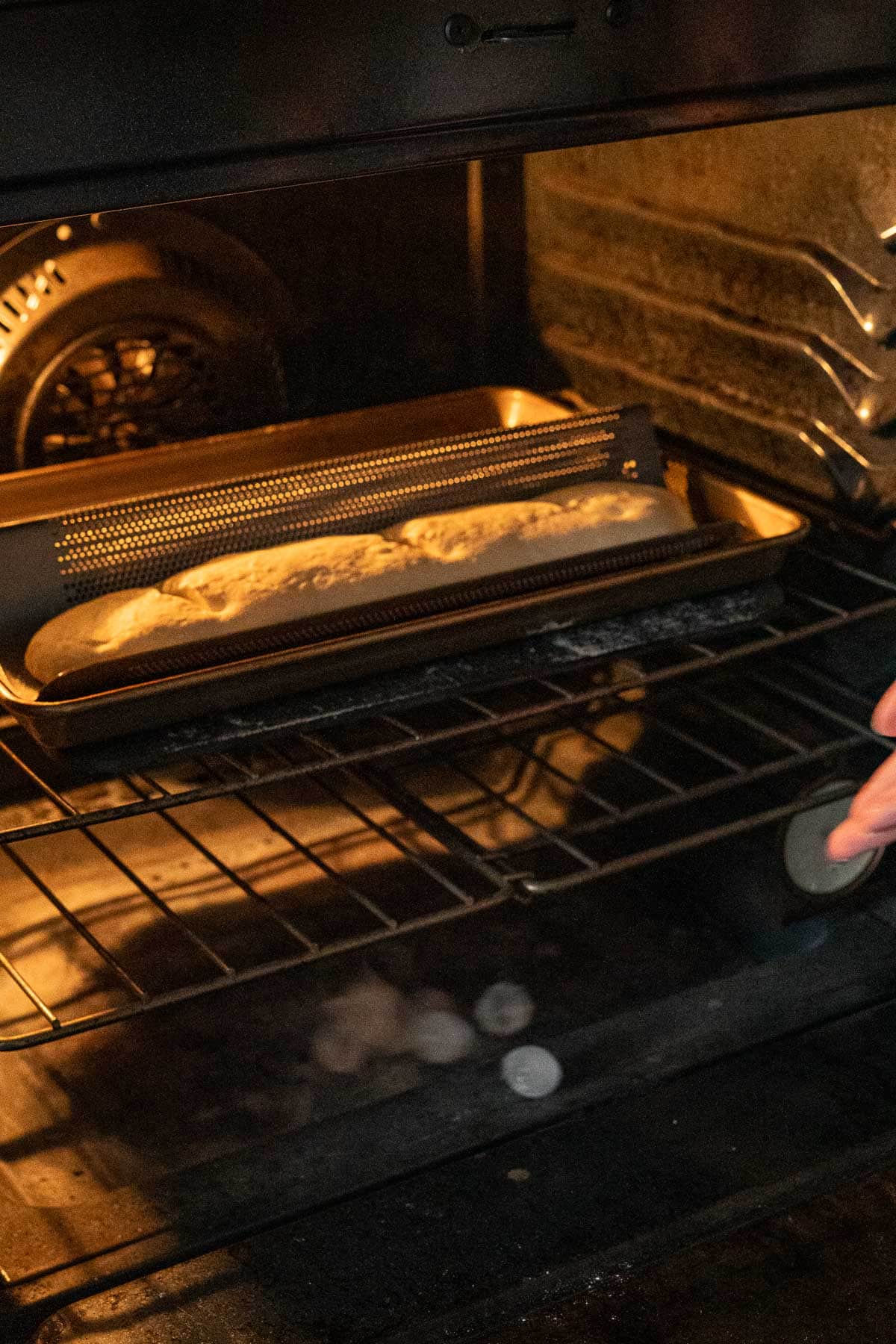 A loaf of unbaked Italian Sourdough Bread dough rests on a slotted baguette pan inside an oven, with a hand steadying the oven rack. The oven’s interior is partially lit and ice cubes are being thrown into the oven.