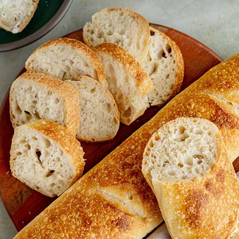 Sliced pieces of Italian Sourdough Bread and a whole baguette are arranged on a round wooden board, highlighting their golden crust and airy, textured interior.