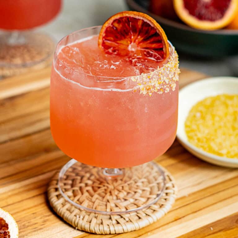 A vibrant blood orange margarita in a glass with ice, garnished with a slice of blood orange on the rim. The rim is partially coated with salt or sugar. The glass sits on a woven coaster on a wooden surface.