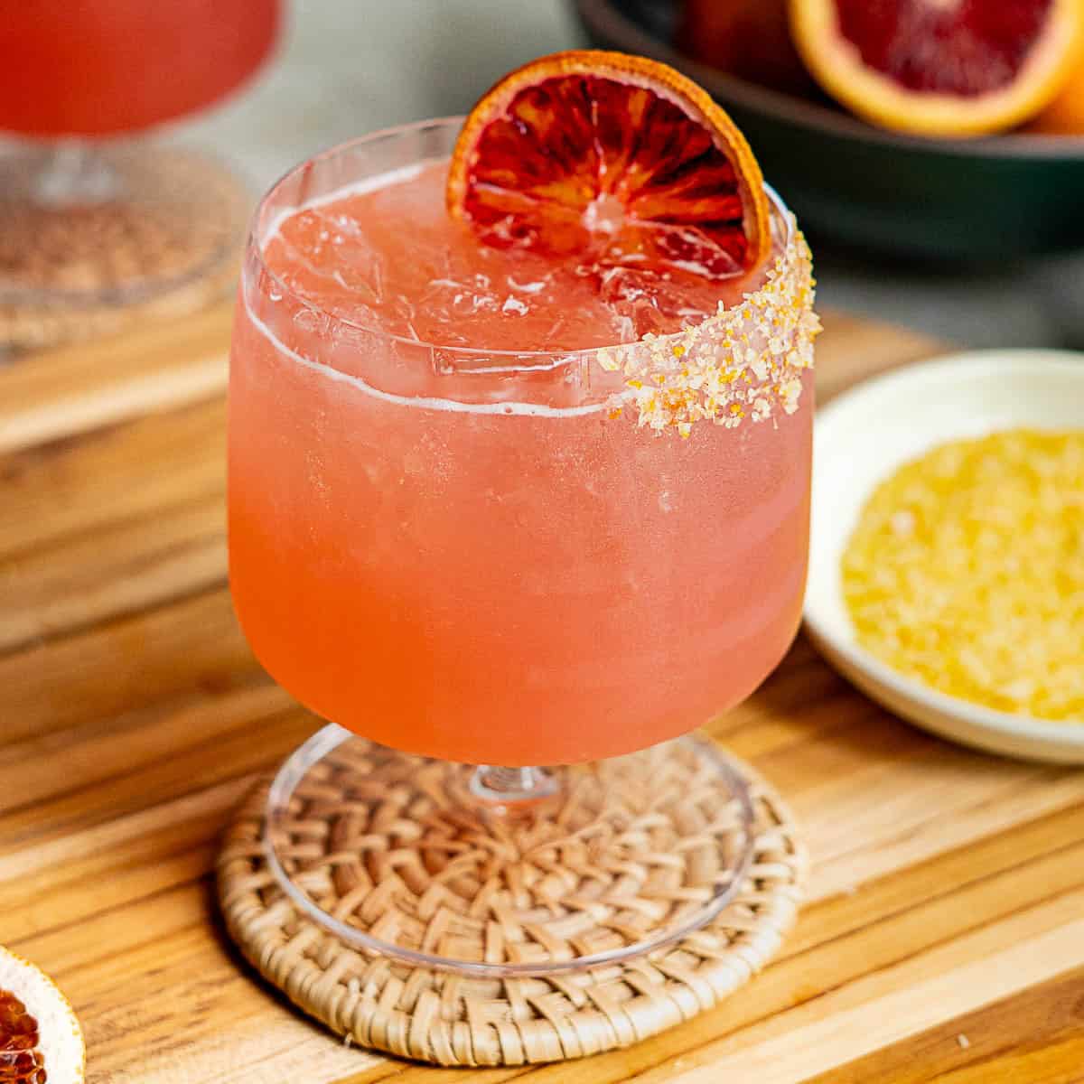 A vibrant blood orange margarita in a glass with ice, garnished with a slice of blood orange on the rim. The rim is partially coated with salt or sugar. The glass sits on a woven coaster on a wooden surface.
