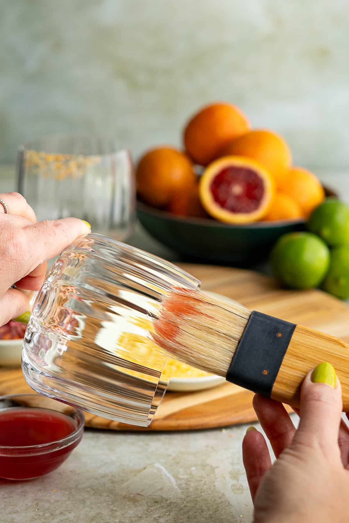 A person brushes the rim of a clear glass with citrus syrup, preparing a blood orange margarita. Surrounded by a bowl of blood oranges and limes, a dish of sugar, and a small bowl of red liquid on a wooden board.