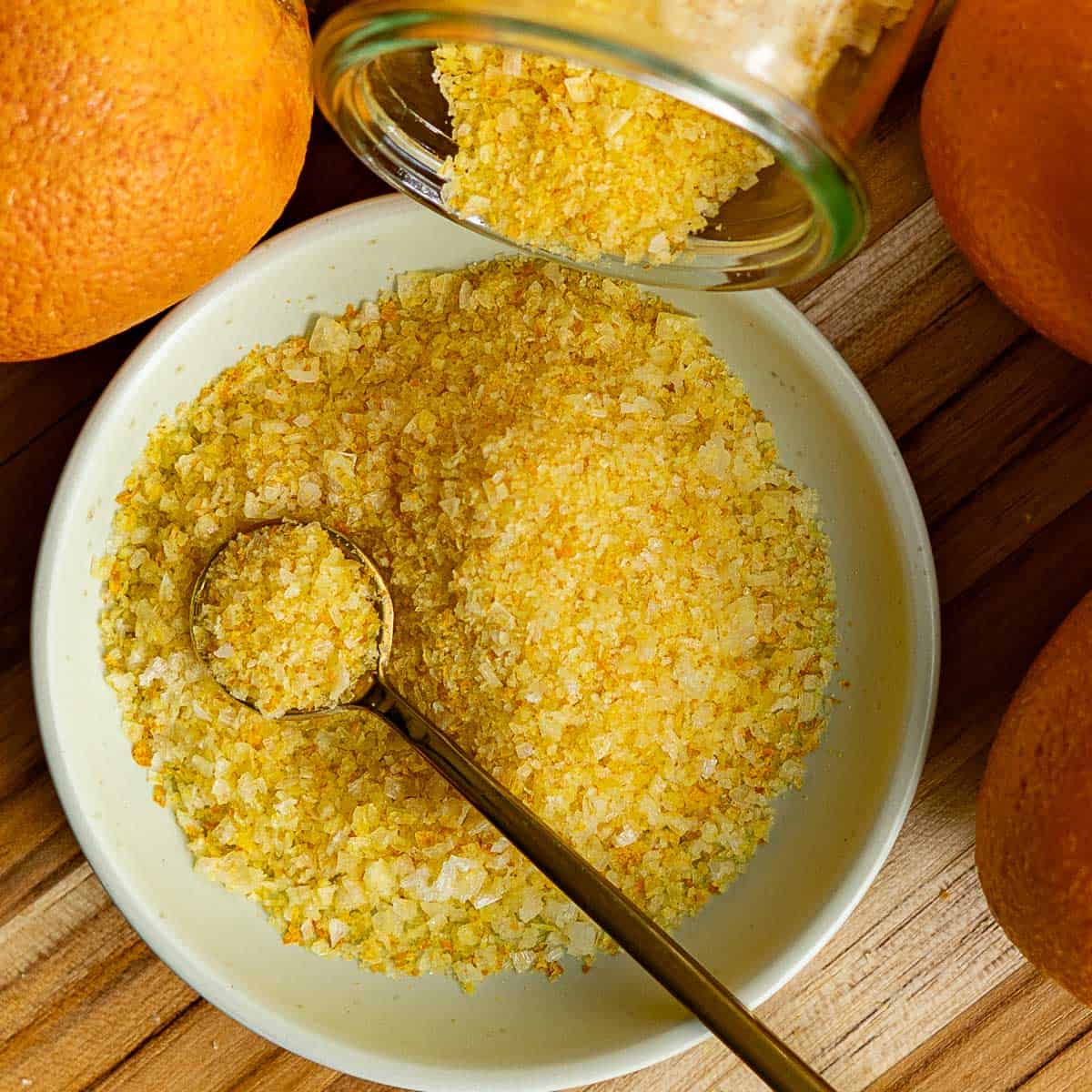 A bowl of orange zest salt with a spoon, surrounded by fresh oranges. A jar is tipped, pouring more orange salt into the bowl on a wooden surface.
