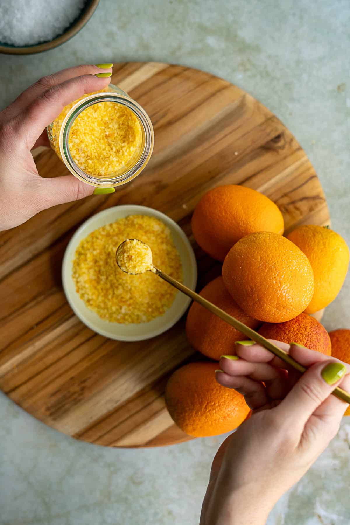 A person with green nail polish spoons yellow-orange citrus salt from a bowl into a small jar, next to whole oranges on a round wooden board.