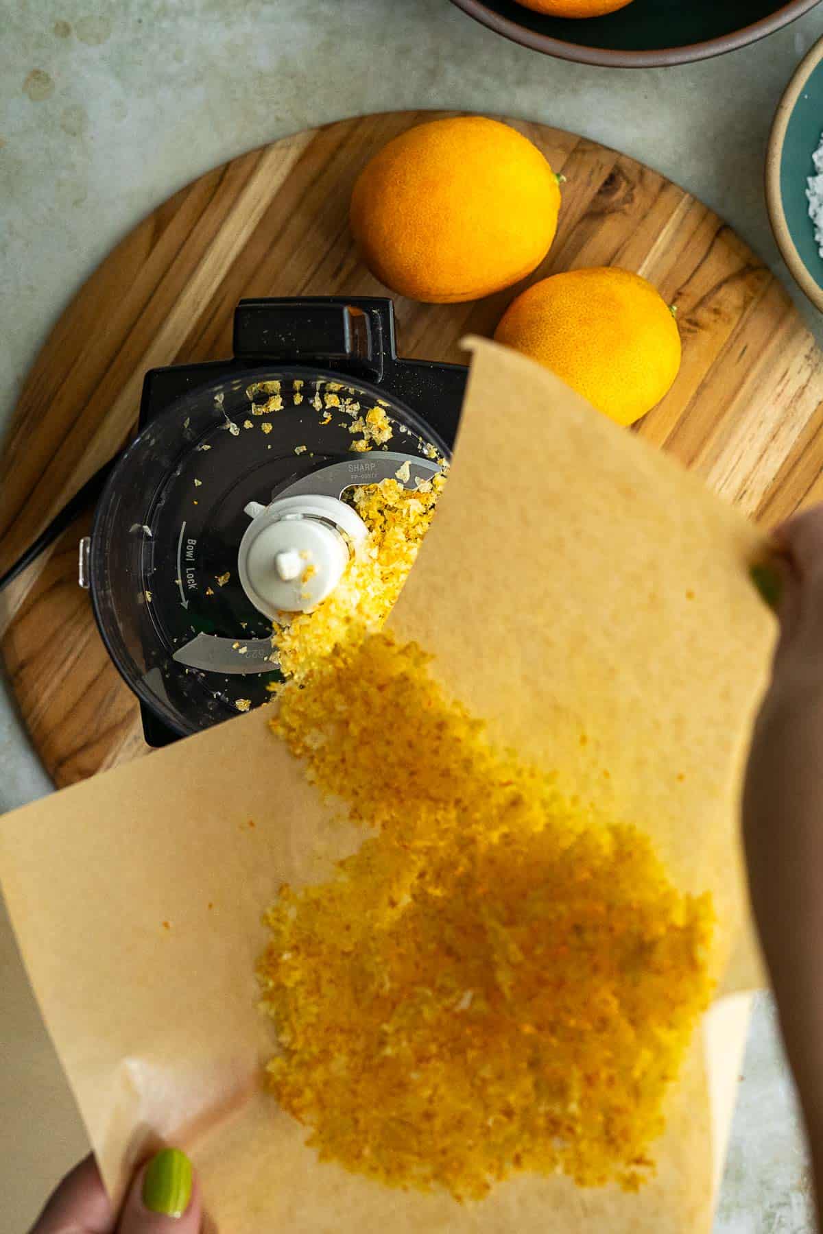 A person pours orange zest salt from parchment paper into a food processor, with two whole oranges and bowls in the background on a wooden board.