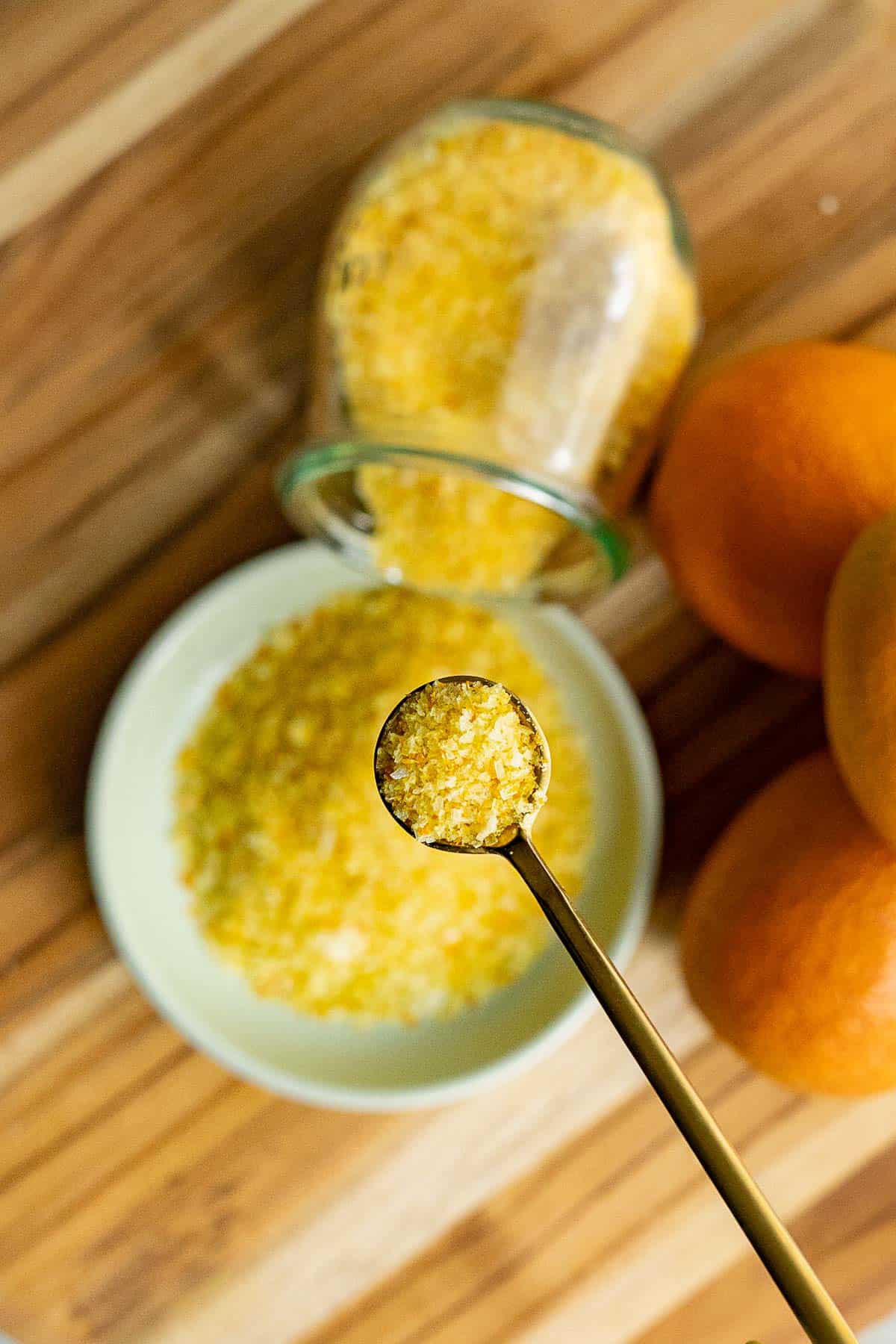 A spoon holds orange salt above a bowl and a jar, both filled with orange zest infused salt. Two whole oranges rest beside them on a wooden surface.