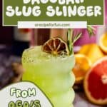 A green, wavy glass filled with a Dagobah Slug Slinger cocktail, garnished with dried lime and rosemary, sits on a table with grapefruit halves and a dark bottle in the background. Text reads, Star Wars Drink—from Oga’s Cantina at home!.