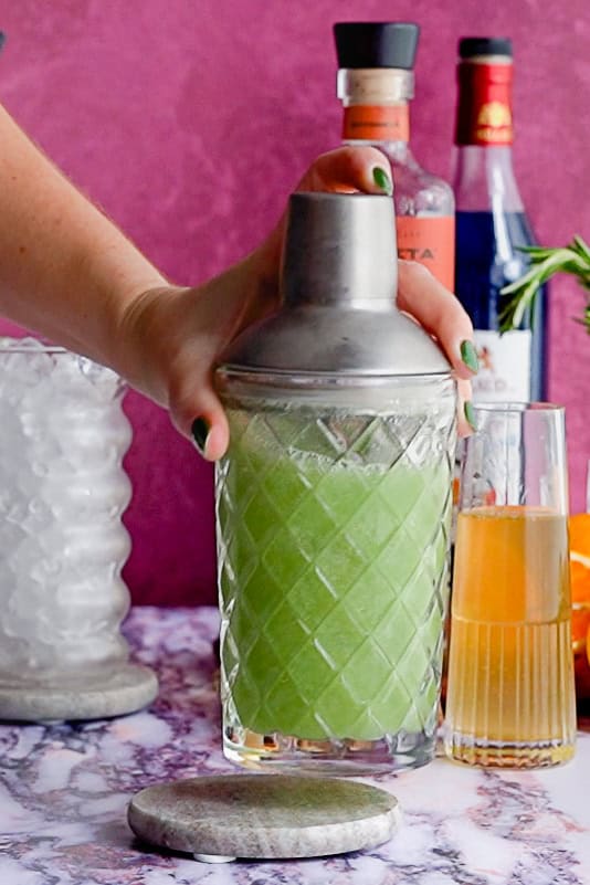 A hand with green-painted nails holds a cocktail shaker filled with a green Dagobah slug slinger. Behind it are bottles, a glass of reposado tequila, a glass of ice, and rosemary, all on a marbled surface against a red background.