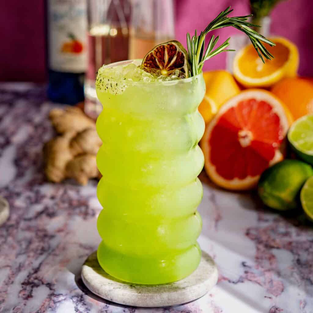 A vibrant green dagobah slug slinger cocktail with ice, garnished with a dried lime slice and rosemary sprig, in a wavy glass on a marble coaster. Citrus fruits, ginger, and a bottle are blurred in the background.