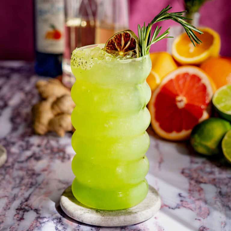 A vibrant green dagobah slug slinger cocktail with ice, garnished with a dried lime slice and rosemary sprig, in a wavy glass on a marble coaster. Citrus fruits, ginger, and a bottle are blurred in the background.