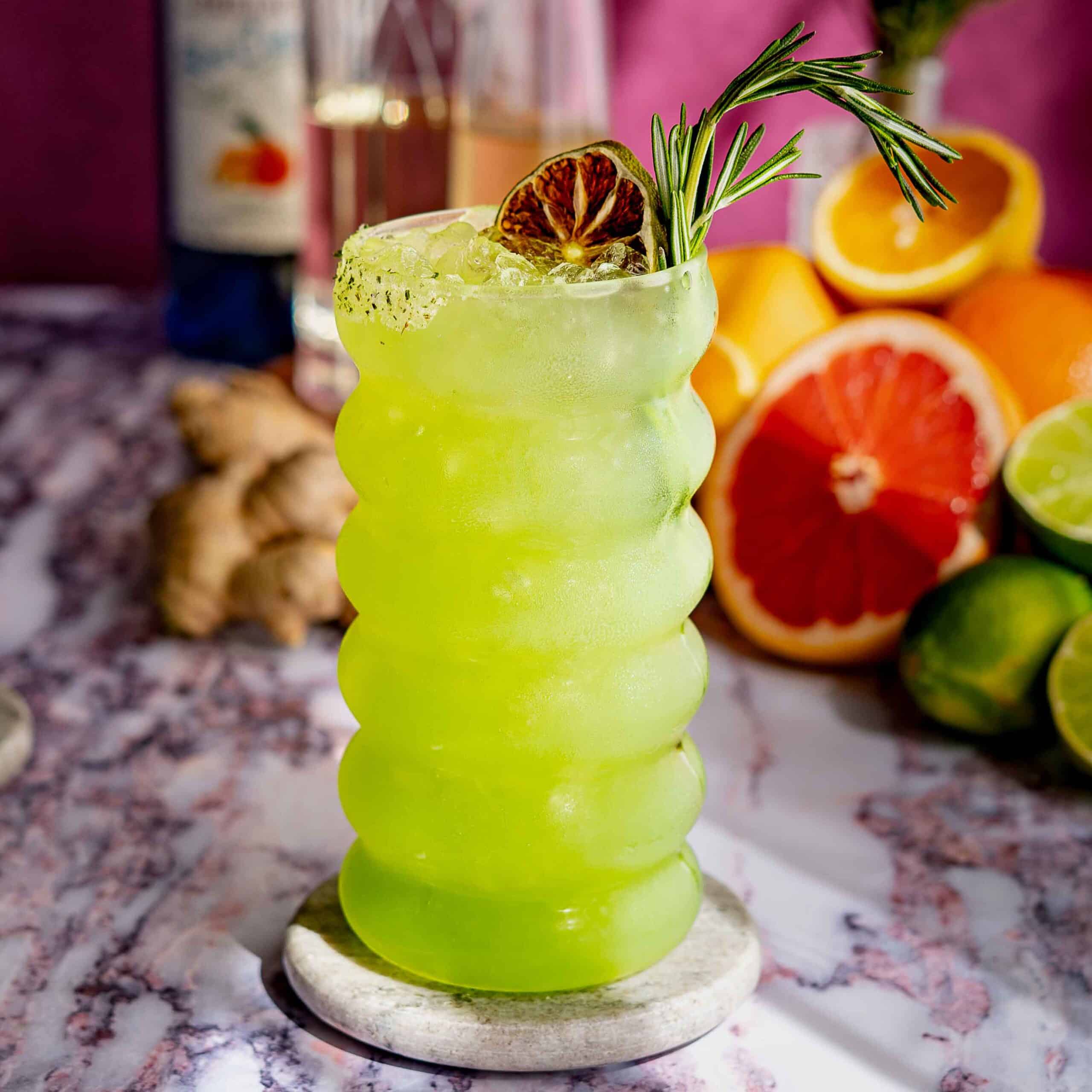 A vibrant green dagobah slug slinger cocktail with ice, garnished with a dried lime slice and rosemary sprig, in a wavy glass on a marble coaster. Citrus fruits, ginger, and a bottle are blurred in the background.