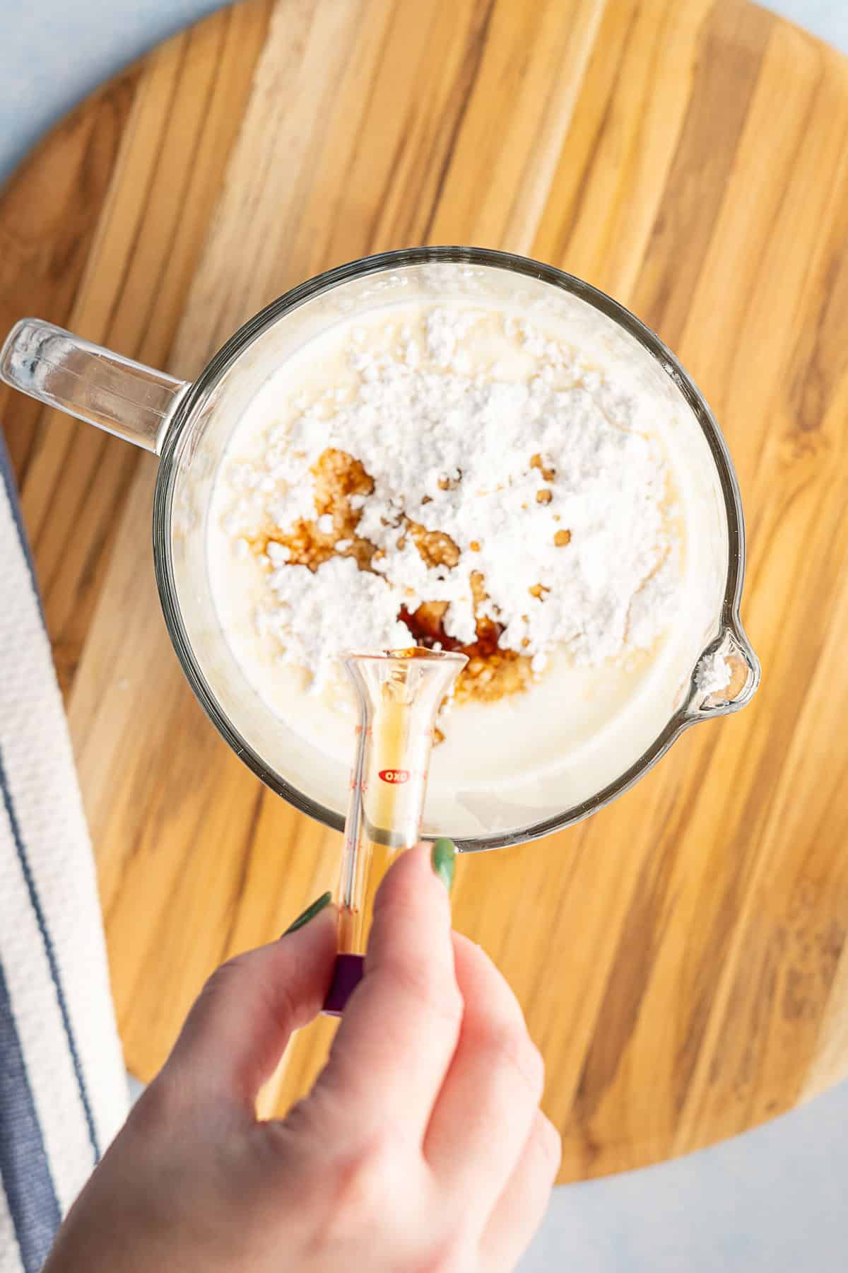 A hand holds a small measuring cup of liquid above a glass bowl containing cream, powdered sugar, and vanilla, all set on a wooden board.