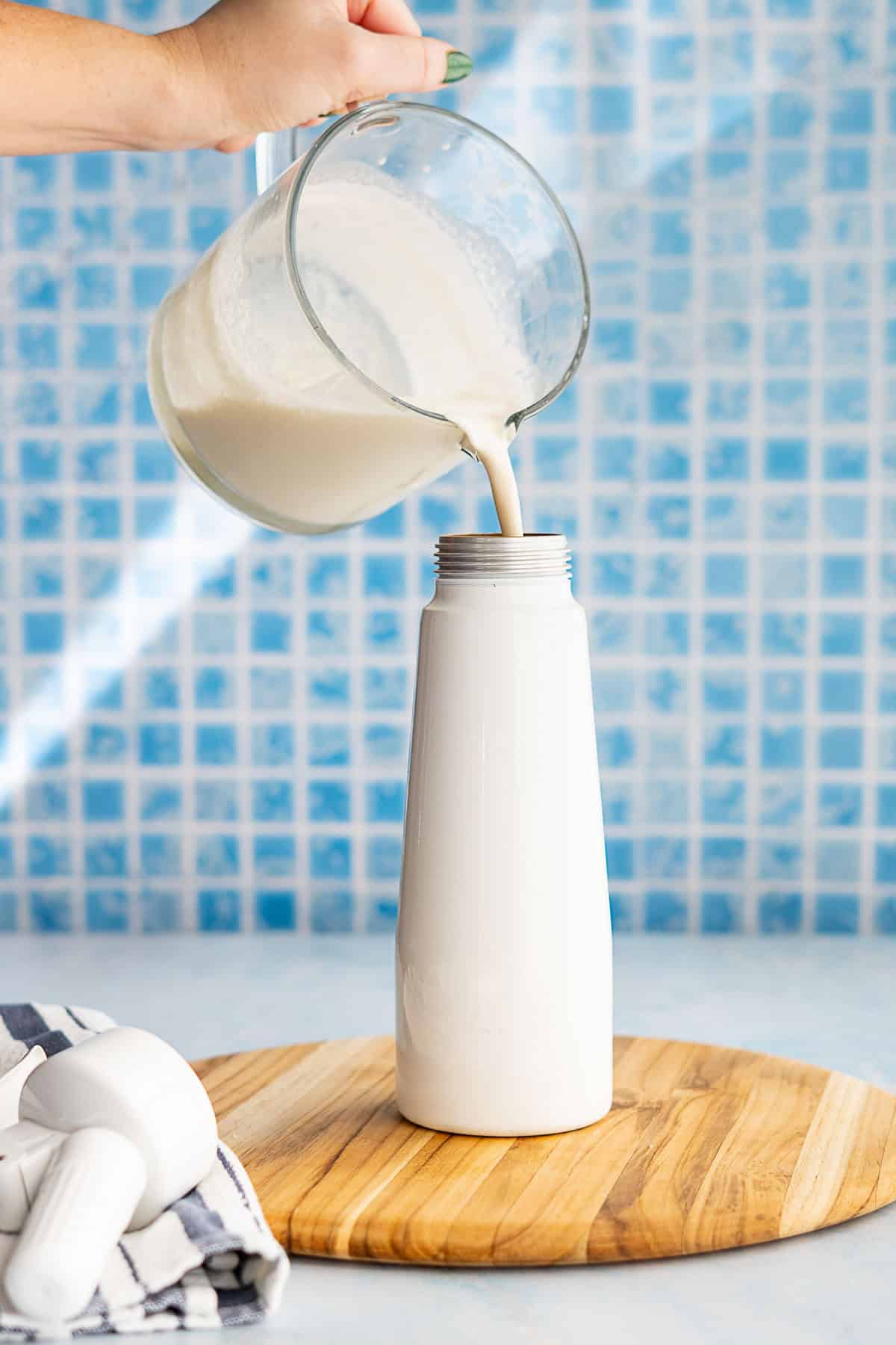 A hand pours a whipped cream base mixture from a glass measuring cup into a whipping canister on a wooden board, with a blue mosaic tile background. A striped cloth and a kitchen tool are visible beside the bottle.