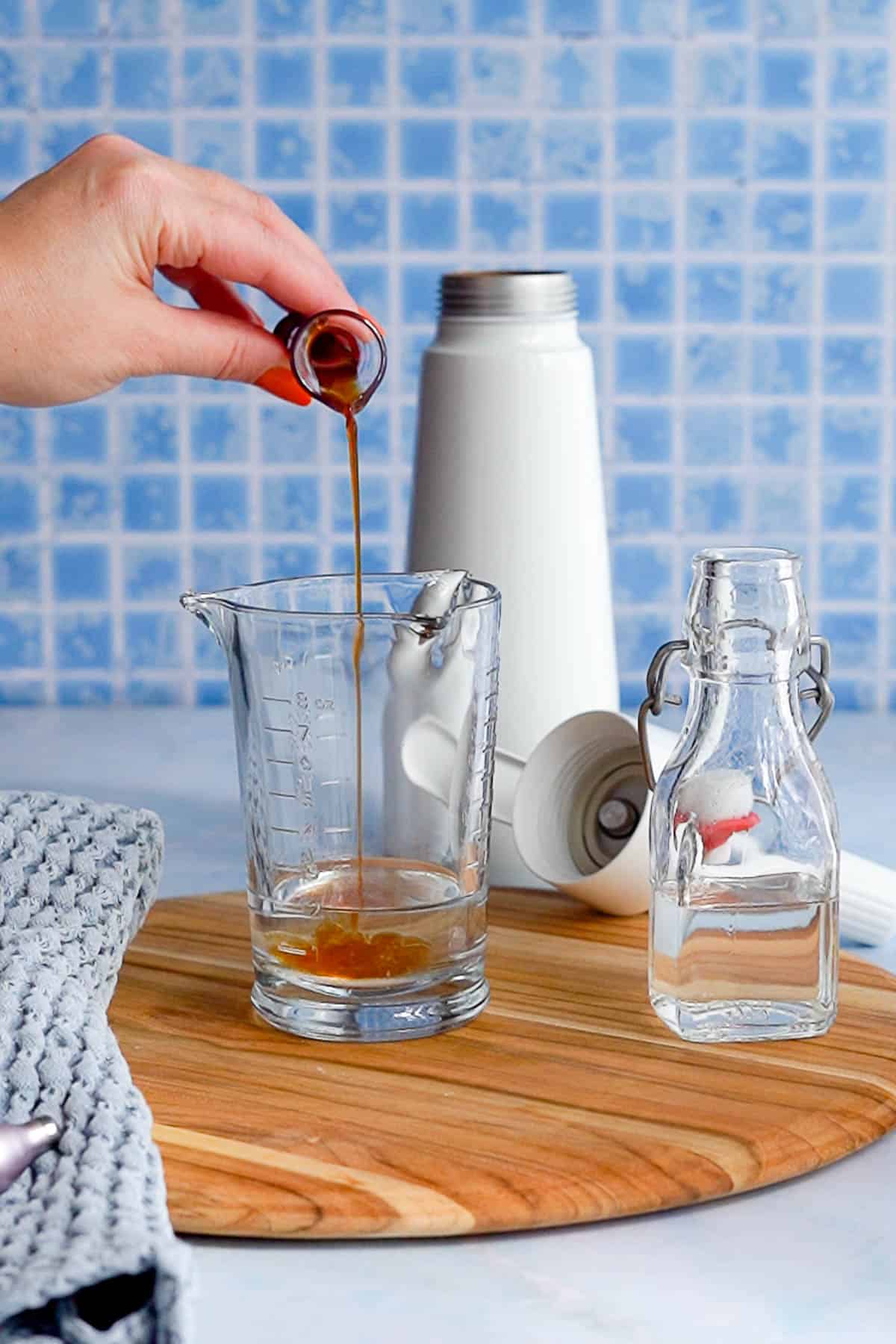 A hand pours vanilla extract from a small container into a glass measuring cup on a wooden board, with a glass bottle, a gray towel, and a white whipped cream dispenser appliance nearby, against a blue tiled background.