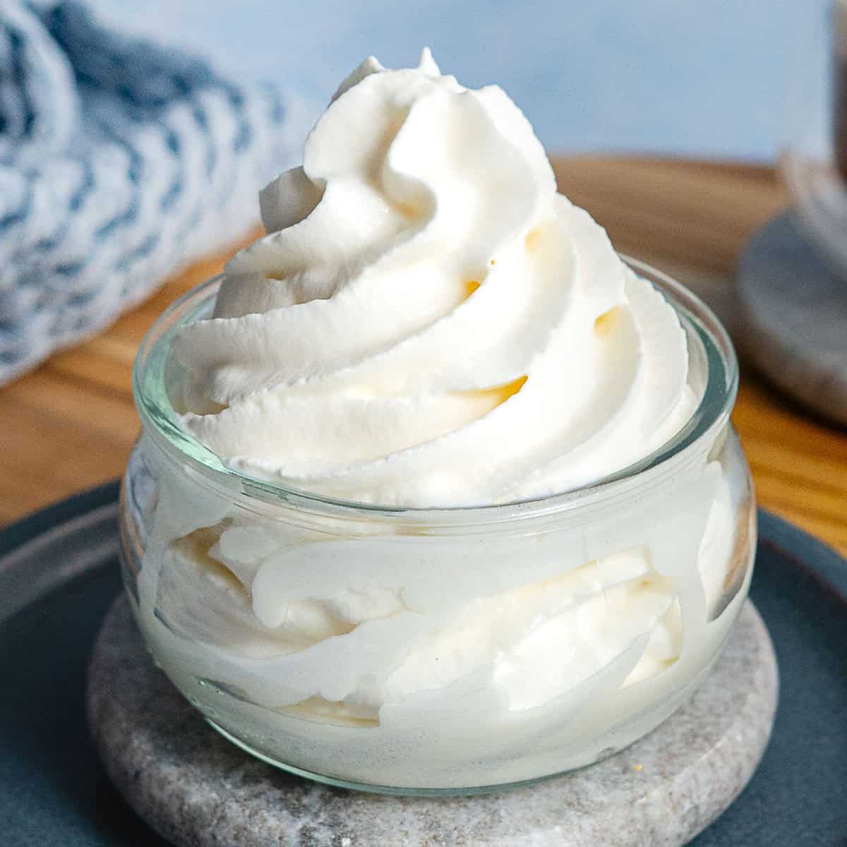 A small glass bowl filled with swirled, fluffy whipped cream made from a whipped cream dispenser sits on a plate with a wooden surface and a textured blue cloth in the background.