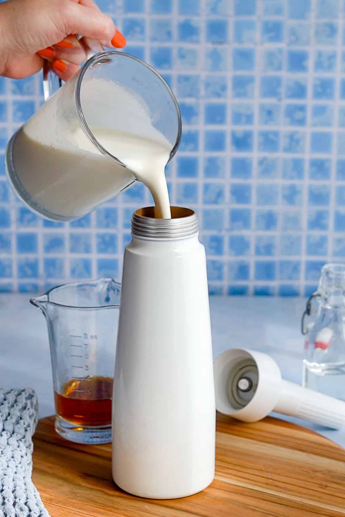 A hand pours a whipped cream base liquid from a measuring cup into a whipped cream siphon on a wooden surface. A glass of amber liquid, an immersion blender, and a blue rag are nearby. The background features blue tile.