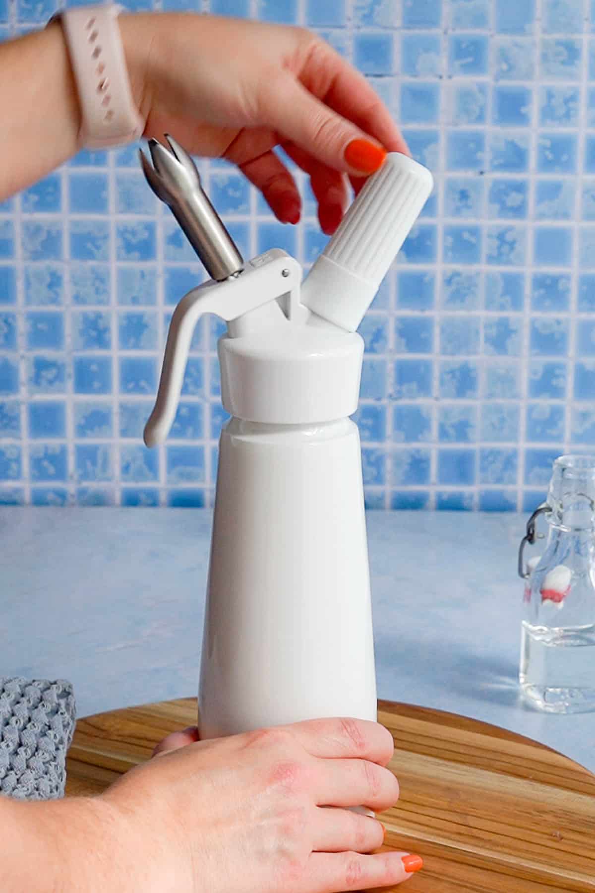 A person’s hands are assembling a white whipped cream dispenser on a wooden board, with a blue tiled wall and a clear glass bottle in the background.