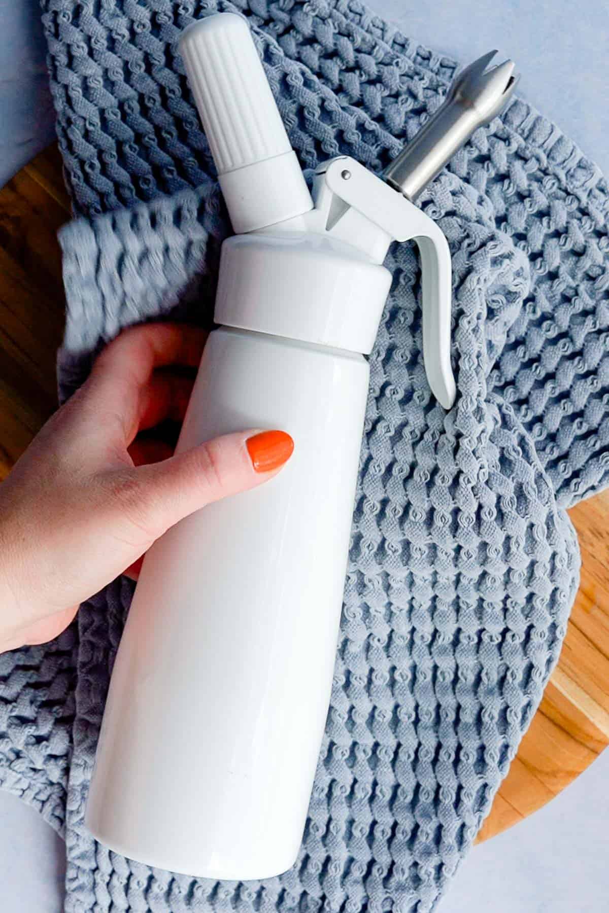 A hand with orange-painted nails holds a white whipped cream dispenser on a textured blue-gray cloth atop a wooden surface.