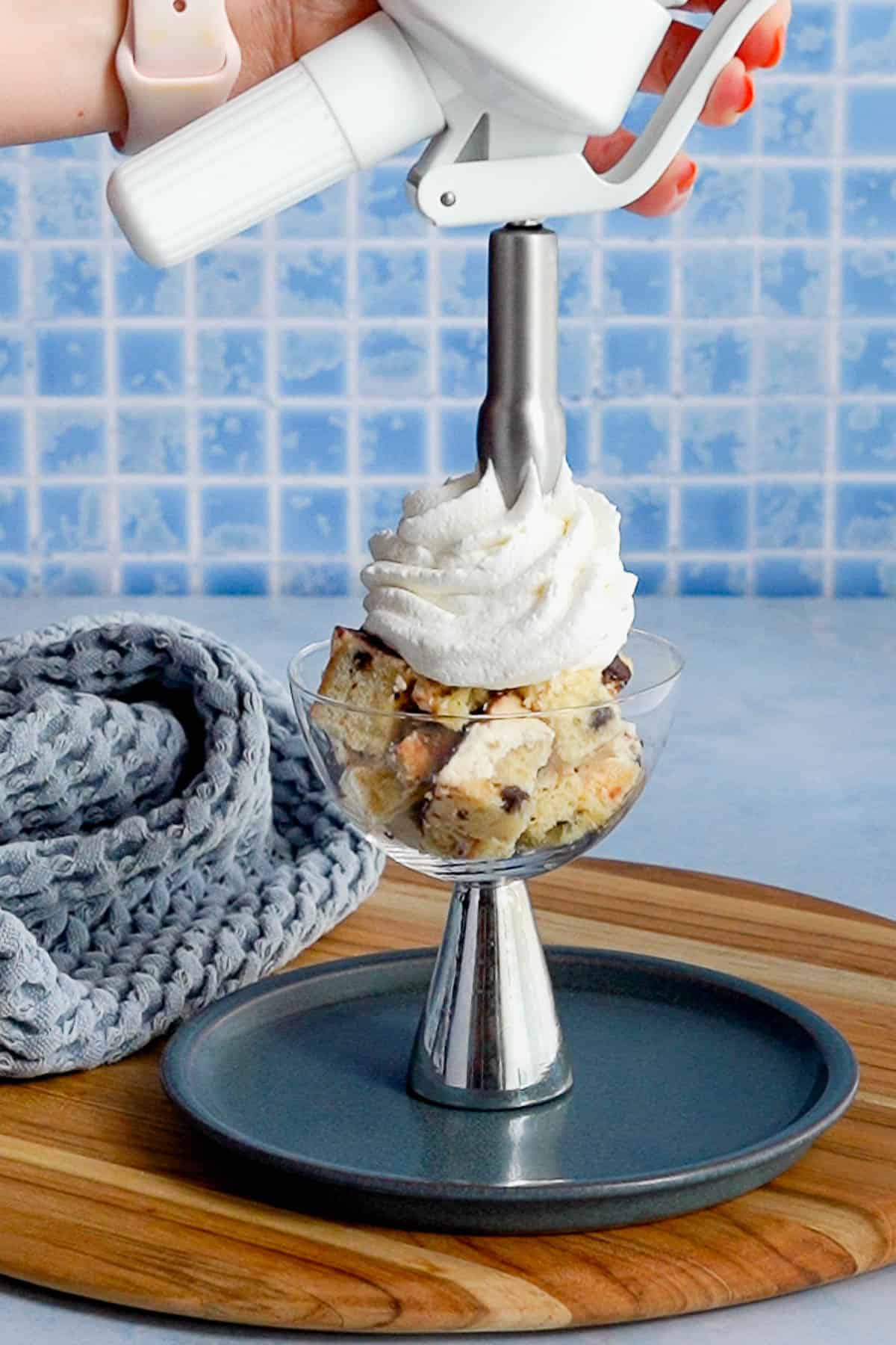 A hand uses a whipped cream dispenser to add a swirl of whipped cream to a dessert glass filled with cookie dough chunks, set on a blue plate with a gray cloth and tiled blue background.