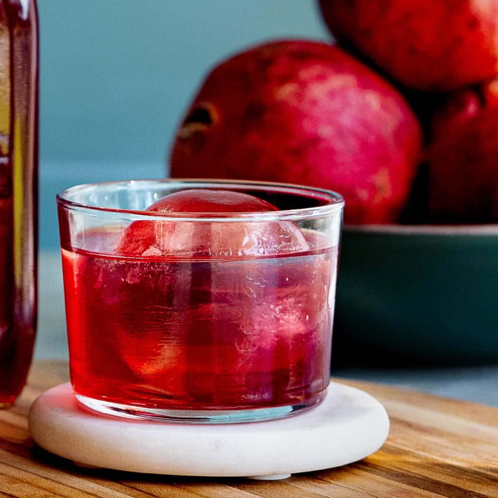 A glass of red pomegranate liqueur with a large round ice cube sits on a coaster next to a bottle of pomegranate liqueur, with a bowl of whole pomegranates in the background on a wooden surface.