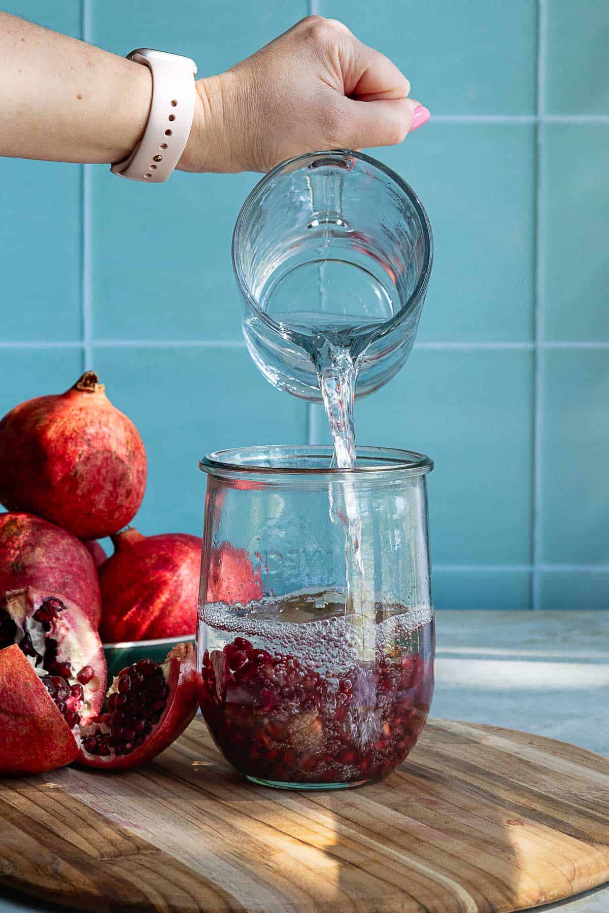 A hand pours vodka from a glass measuring cup into a jar filled with pomegranate seeds, preparing for homemade pomegranate liqueur. Whole and cut pomegranates rest on a wooden board against a backdrop of blue tiles.