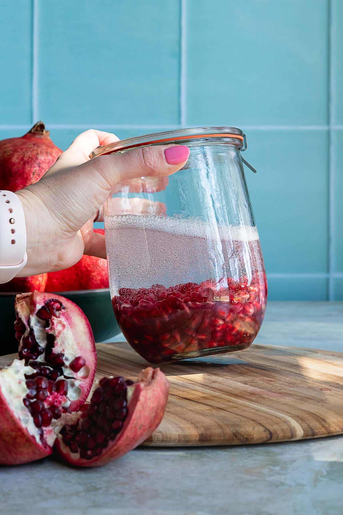 A hand with pink nail polish holds a glass jar filled with pomegranate liqueur and seeds. A whole pomegranate and a pomegranate half rest on a wooden board in front of a blue tiled background.