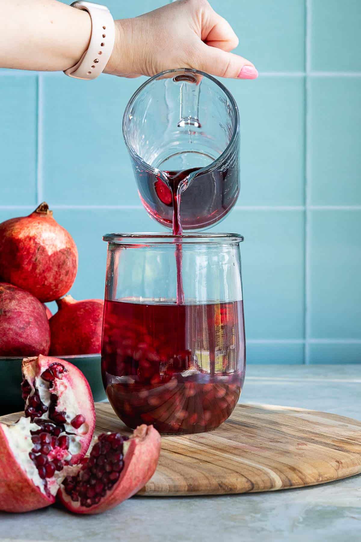 A hand pours pomegranate syrup from a glass into a jar filled with juice and pomegranate seeds, as whole and cut pomegranates and a bottle of pomegranate liqueur rest on a wooden board against a blue tiled background.