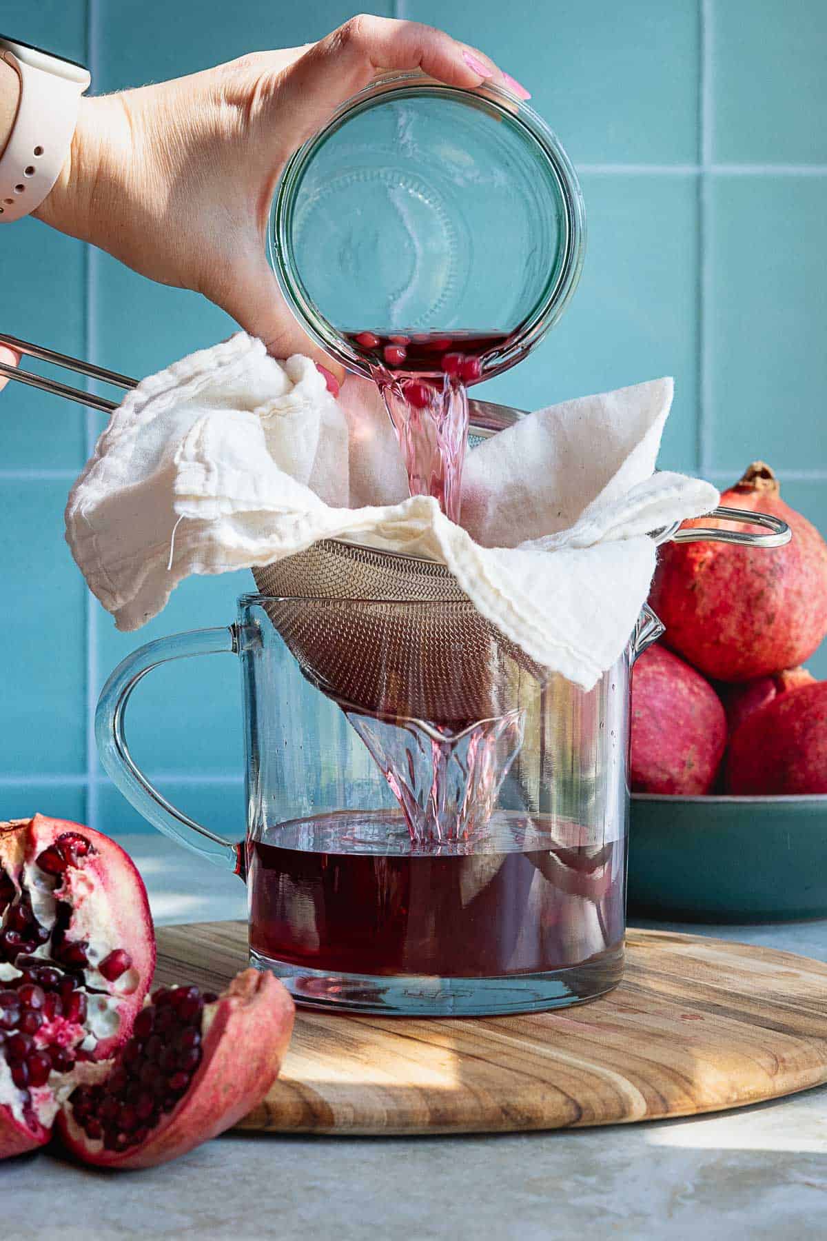 A hand pours pomegranate liqueur through a cloth-lined strainer into a glass container, perfect for crafting homemade pomegranate liqueur, with fresh pomegranates and a wooden cutting board nearby against a blue tiled background.