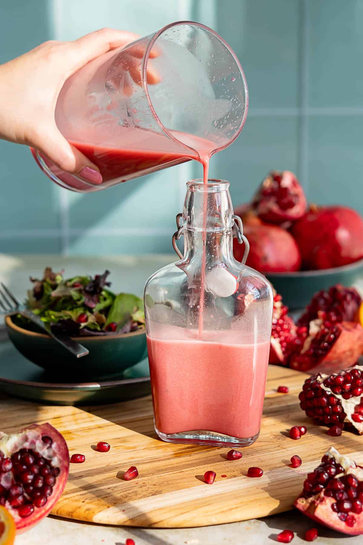 A hand pours red pomegranate salad dressing from a glass measuring cup into a clear glass bottle on a wooden board, surrounded by pomegranate fruits, seeds, and a bowl of salad.