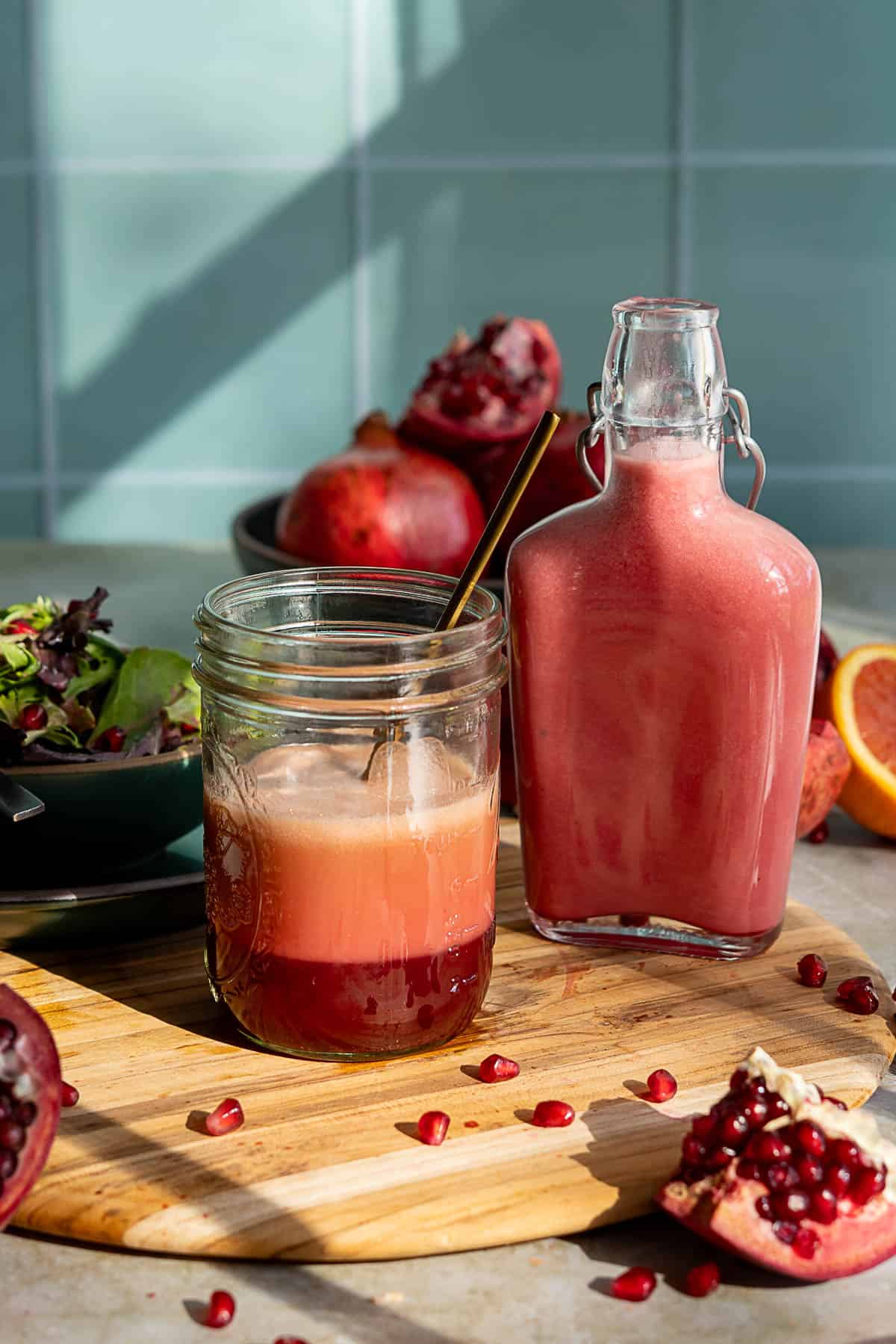 A glass jar holds a broken pomegranate vinaigrette salad dressing beside a matching bottle on a wooden board. Pomegranate seeds, salad, and whole pomegranates are in the background, all lit by natural sunlight.