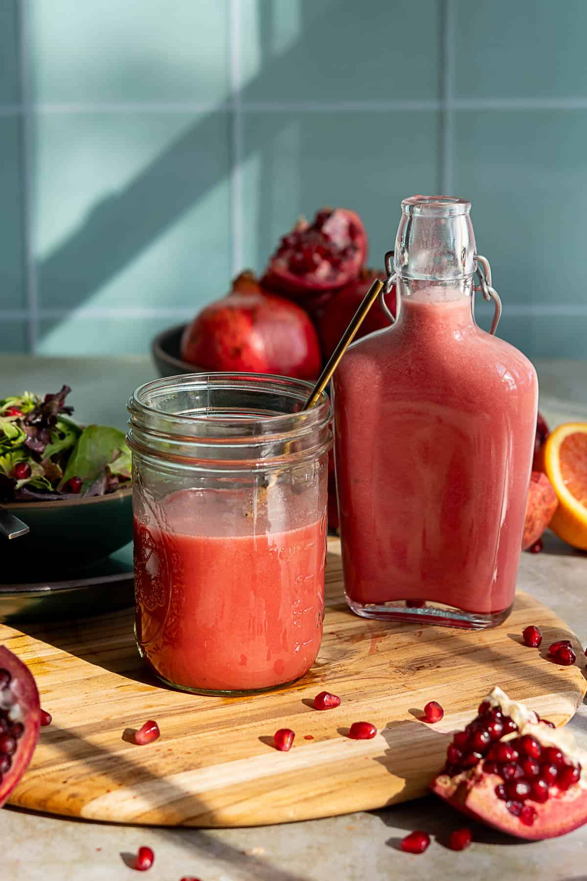 A glass jar holds a properly blended pomegranate salad dressing beside a matching bottle on a wooden board. Pomegranate seeds, salad, and whole pomegranates are in the background, all lit by natural sunlight.