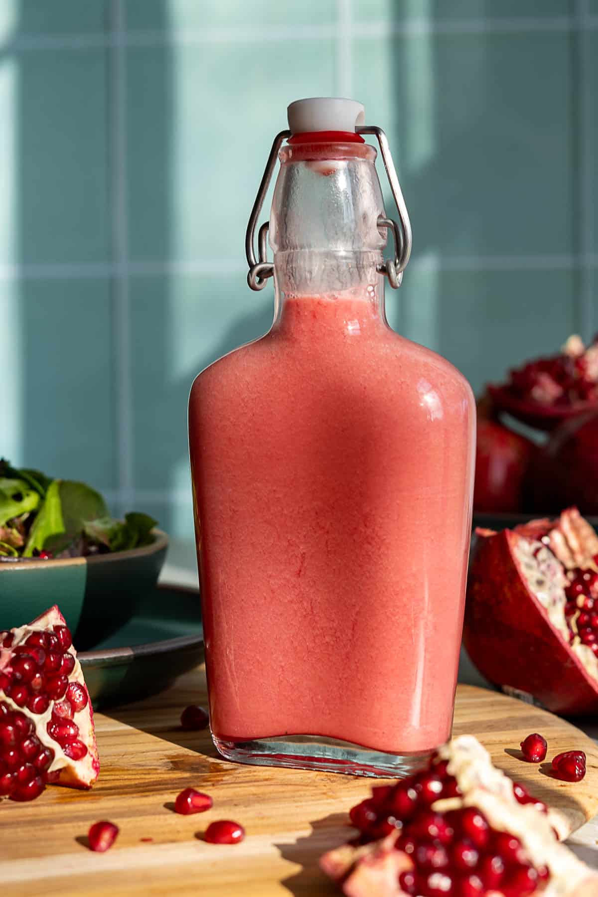 A glass bottle filled with pink pomegranate salad dressing sits on a wooden surface, surrounded by pomegranate halves, seeds, and a bowl of salad, with sunlight casting shadows in the background.
