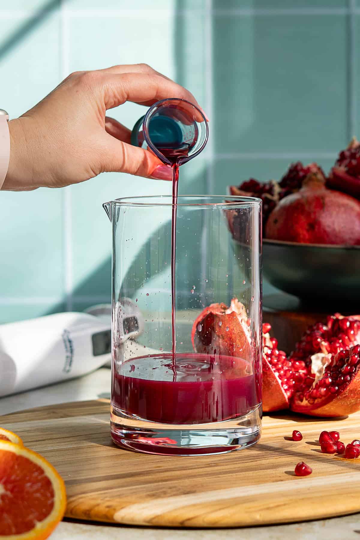A hand pours pomegranate syrup from a small glass into a larger glass beaker on a wooden board, with pomegranate fruit and seeds in the background.