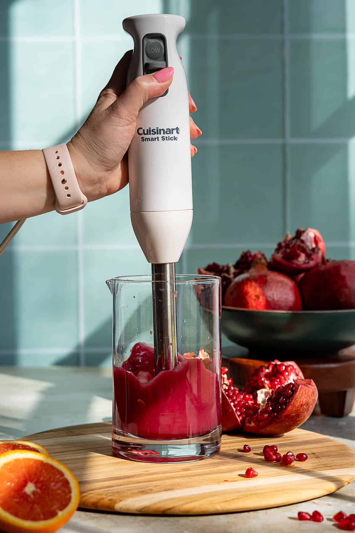 A hand holds a Cuisinart Smart Stick immersion blender blending pomegranate seeds in a glass container, with whole and cut pomegranates and an orange on a wooden board nearby.