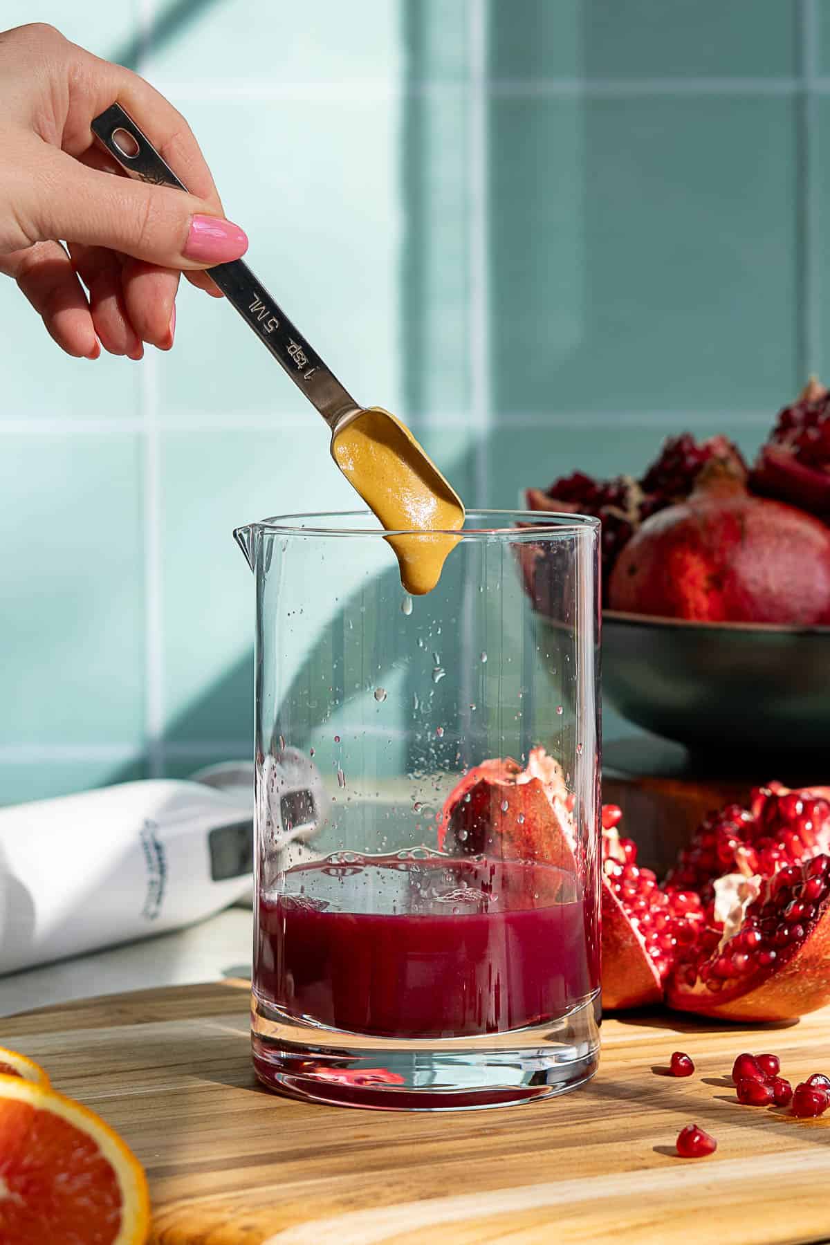 A hand holds a measuring spoon of mustard over a glass filled with pomegranate juice. Cut pomegranates, seeds, and an orange rest on a wooden board, with whole pomegranates in a bowl in the background.