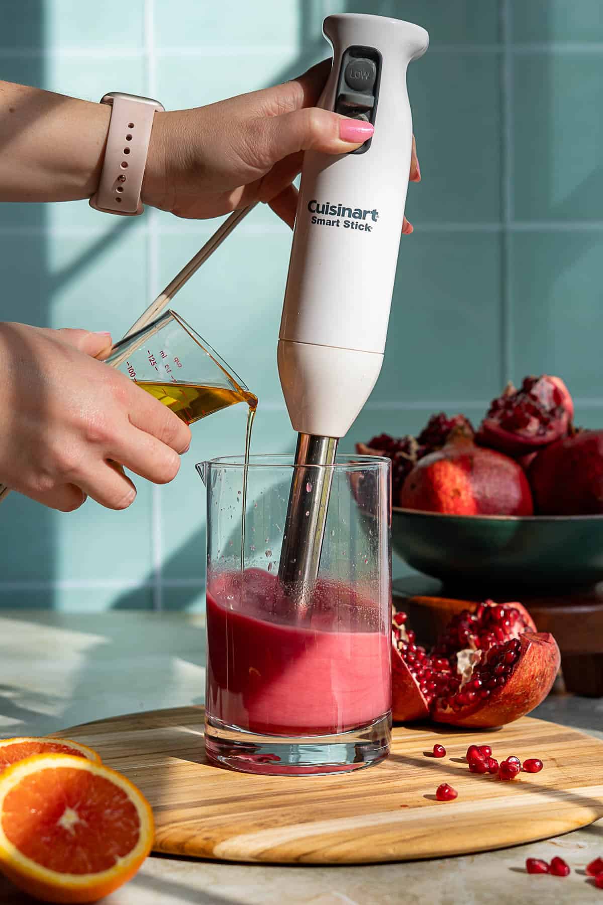 A person uses a Cuisinart Smart Stick hand blender to mix a pomegranate vinaigrette in a glass while pouring liquid from a measuring cup; fresh pomegranates and an orange sit nearby on a cutting board.