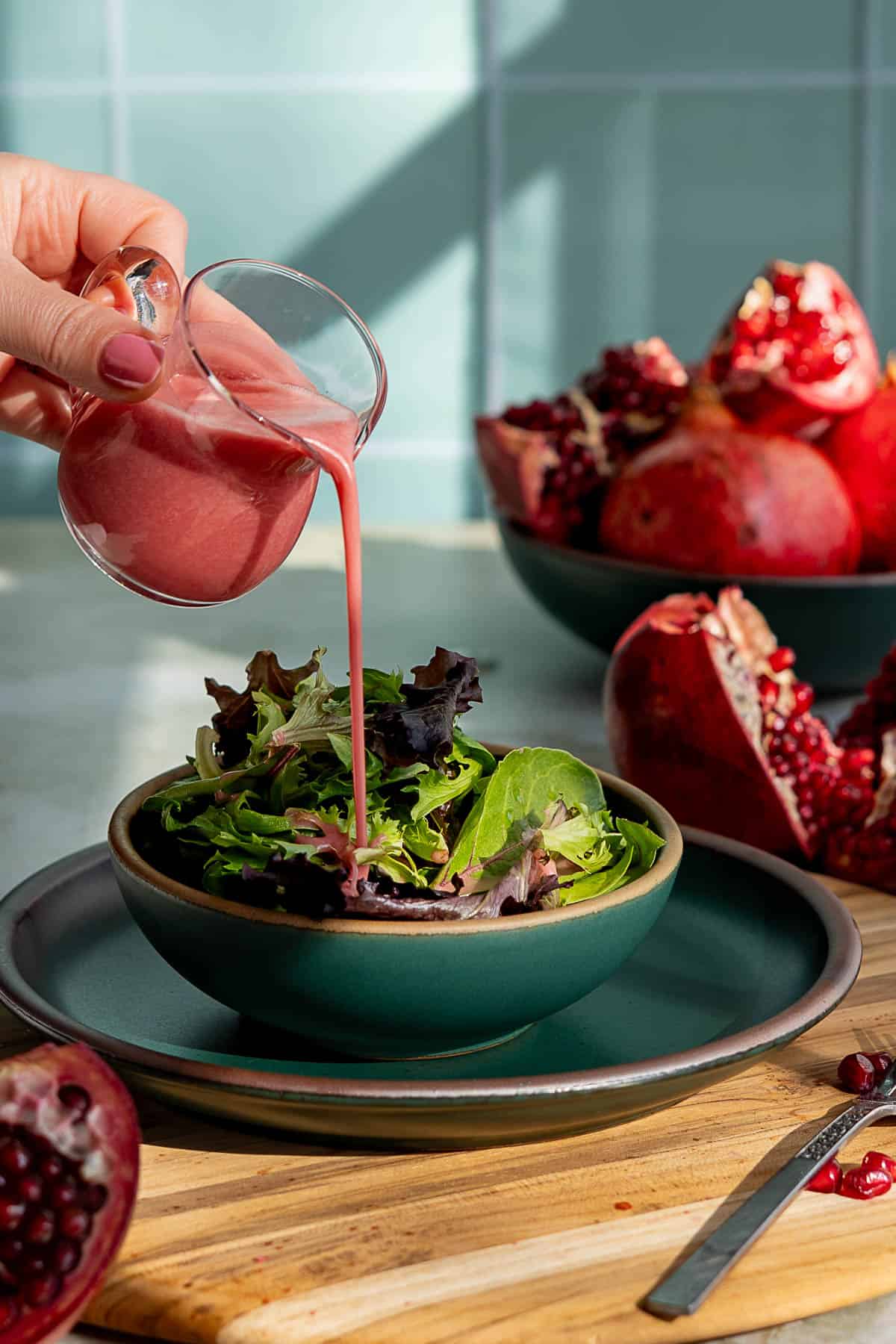 A hand pours pomegranate dressing from a small glass pitcher over a bowl of mixed leafy greens. Sliced pomegranates rest on a wooden board and in a bowl in the background.