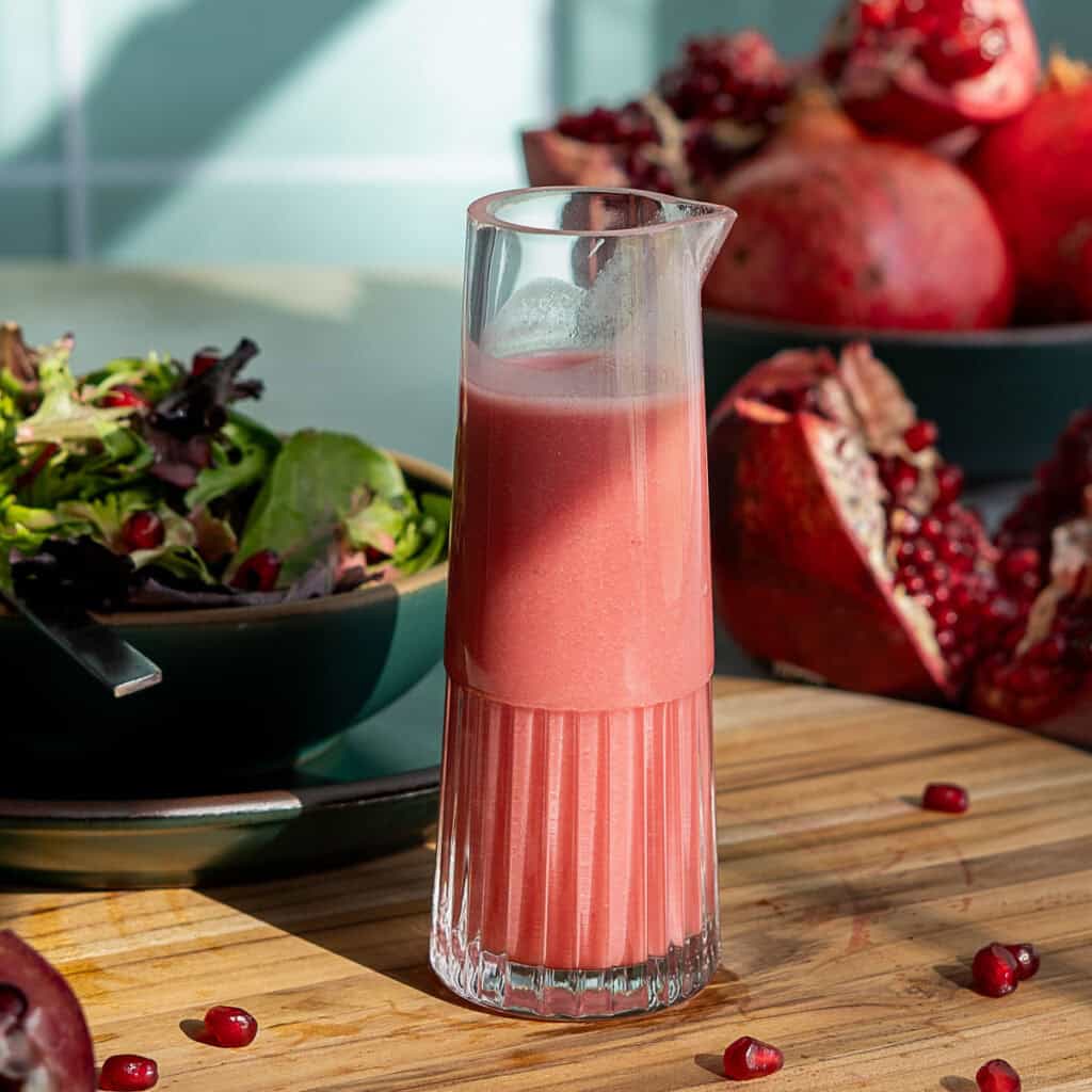A glass pitcher filled with pomegranate salad dressing sits on a wooden surface. In the background, there is a bowl of mixed greens and whole as well as cut pomegranates, with scattered pomegranate seeds around.