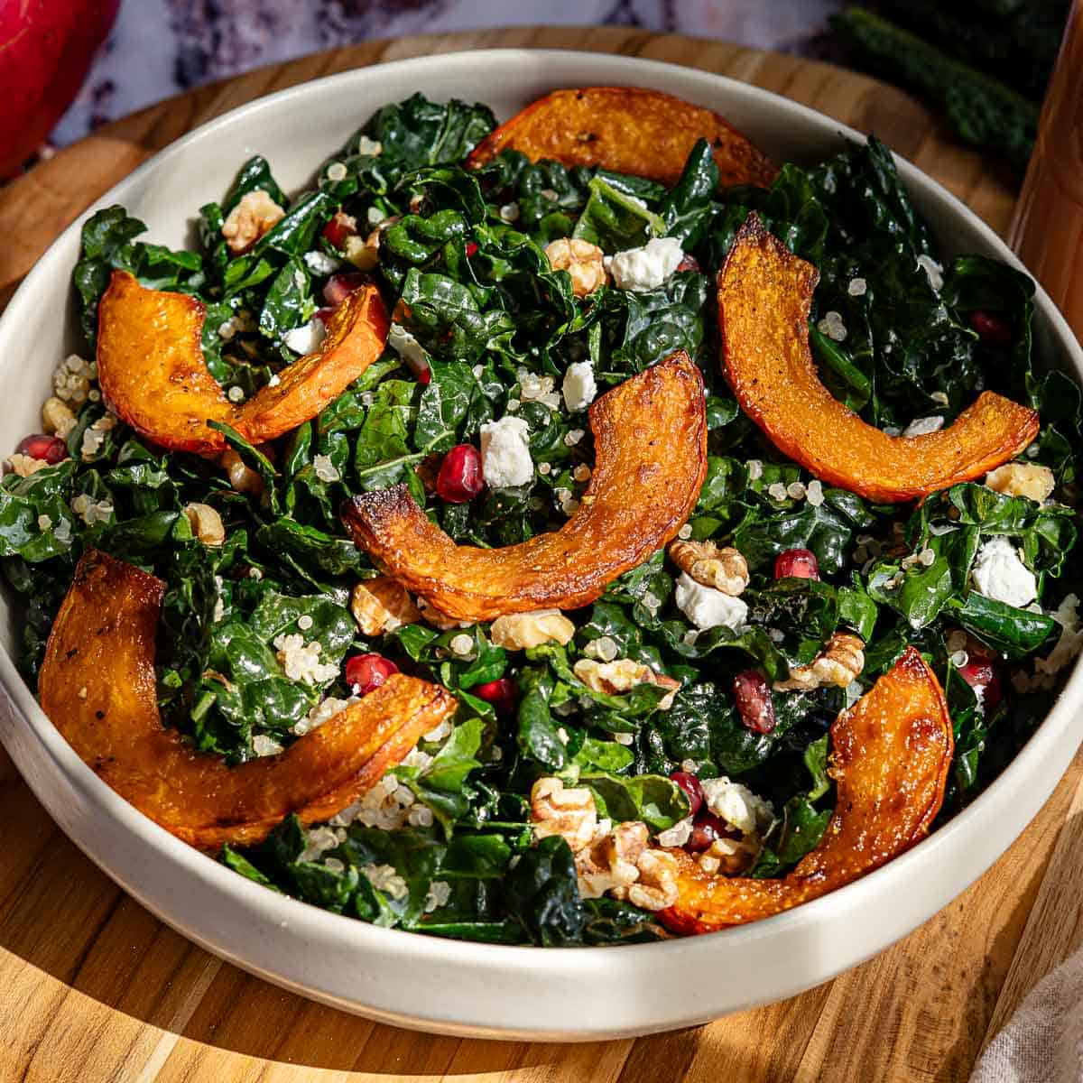 A bowl of a kale salad topped with pomegranate seeds, roasted red kuri squash slices, crumbled feta cheese, walnuts, and quinoa, served on a wooden surface.