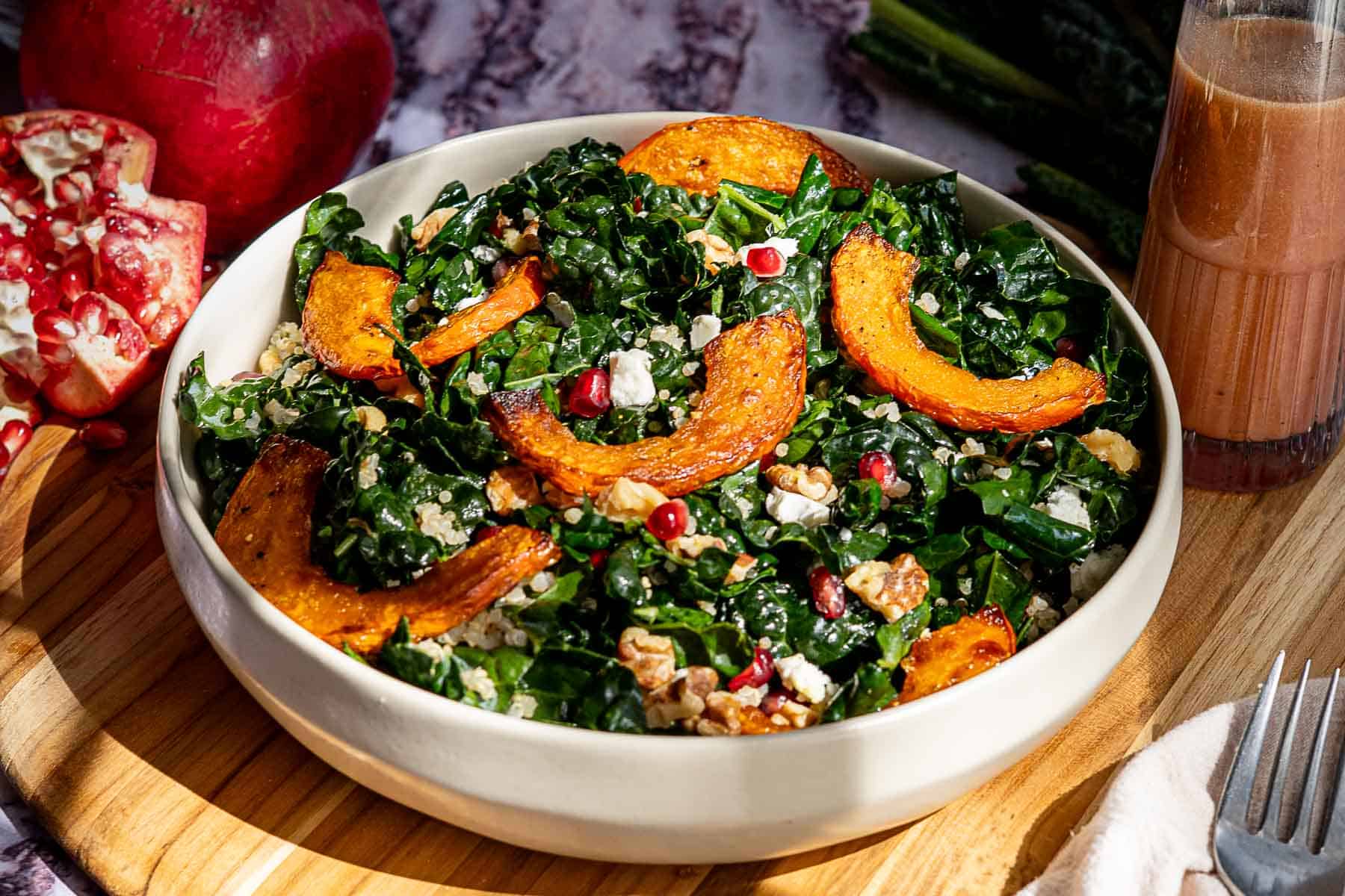 A bowl of kale and pomegranate salad with roasted squash slices, walnuts, feta cheese, and pomegranate seeds sits on a wooden board, beside a halved pomegranate and a glass of dressing.