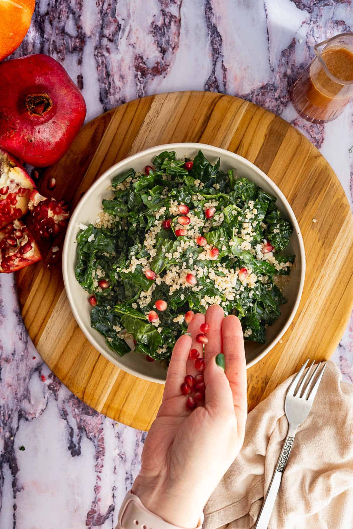 A hand sprinkles pomegranate seeds over a vibrant kale and pomegranate salad with quinoa, set on a round wooden board. Pomegranate halves, a jar of dressing, and a fork rest nearby on the marble surface.