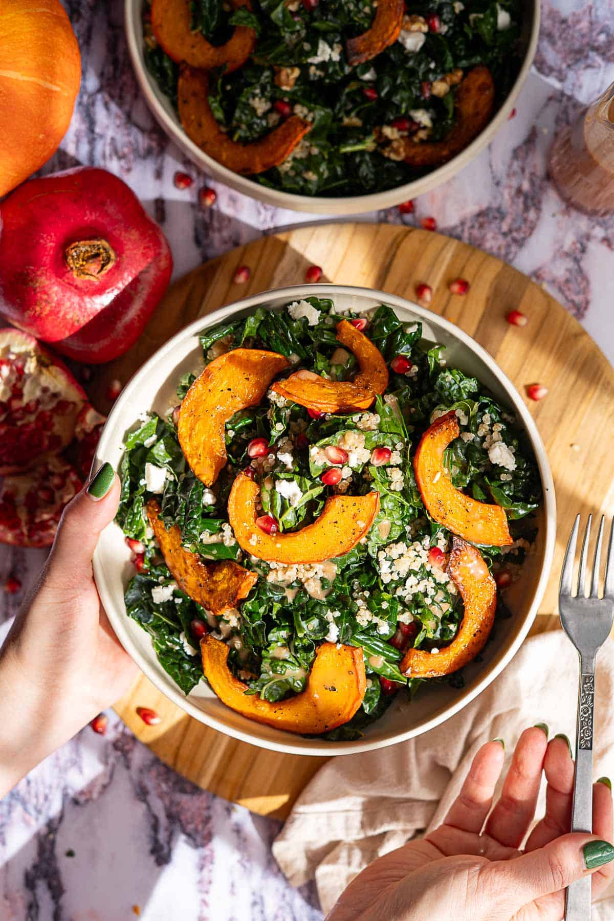 A vibrant kale and pomegranate salad is held above a wooden board, featuring roasted squash slices, grains, and pomegranate seeds. A fork and whole pomegranates sit nearby, with another salad bowl partially visible at the top.