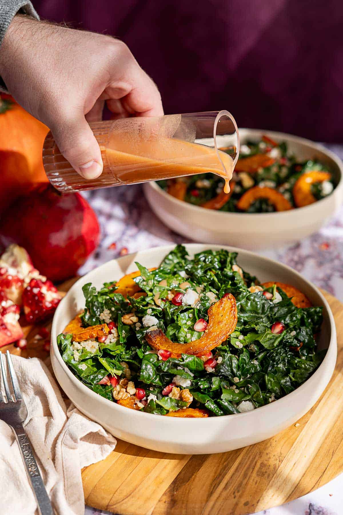 A hand pours salad dressing onto a vibrant kale and pomegranate salad with roasted squash and walnuts. In the background, another bowl of salad, a pomegranate, and a pumpkin sit on a wooden surface.