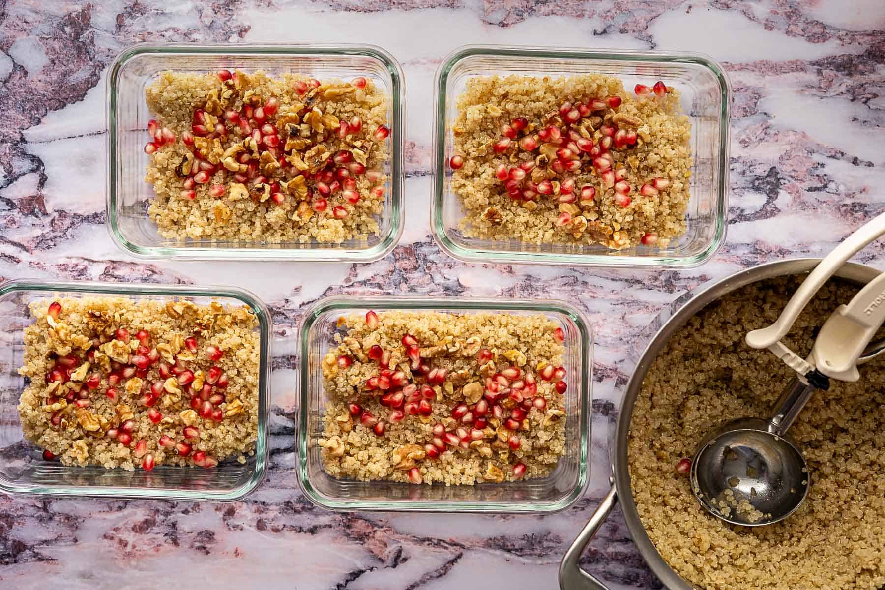 Four glass meal prep containers filled with cooked quinoa, pomegranate seeds, and chopped nuts are arranged on a marble countertop, perfect for pairing with a fresh kale and pomegranate salad. Nearby, a pot with leftover quinoa and a serving scoop waits.