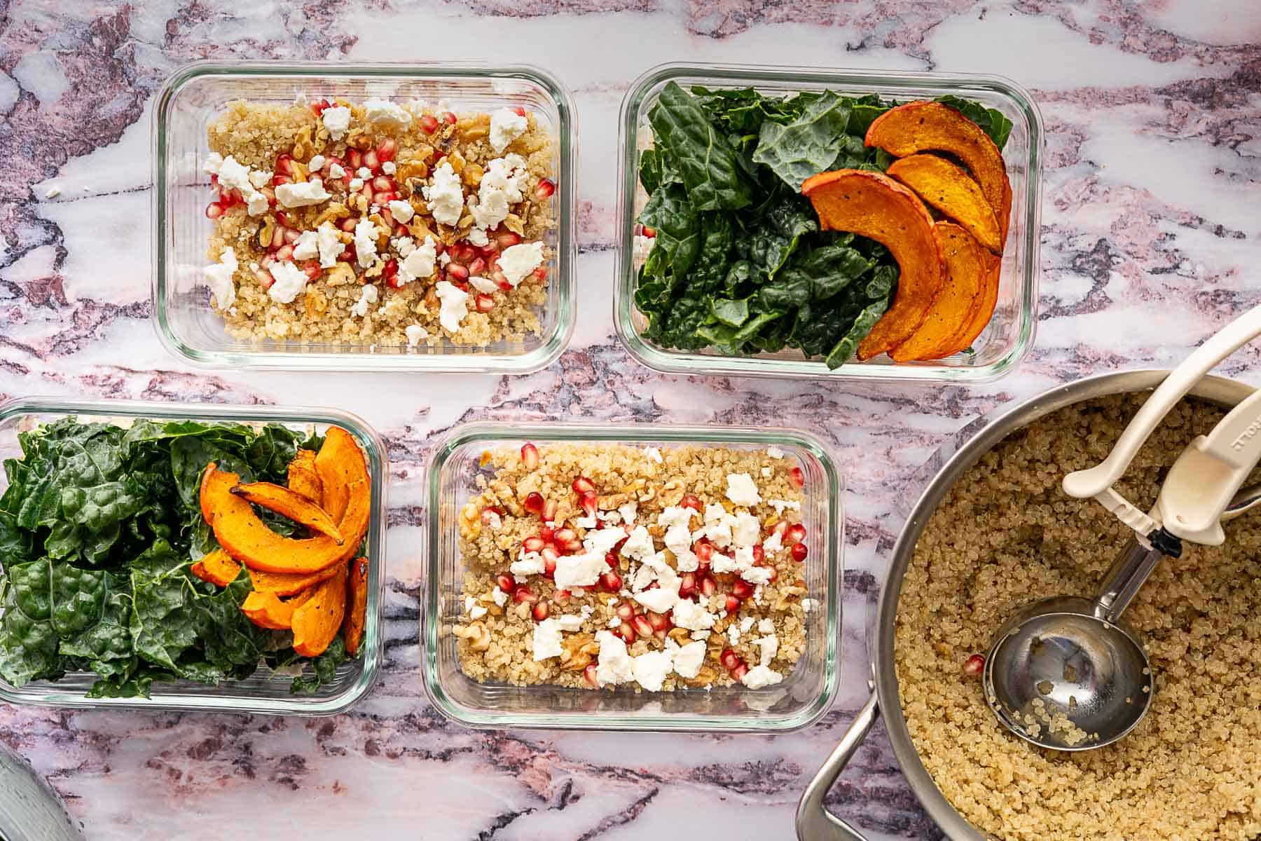 Four glass meal prep containers with quinoa, kale and pomegranate salad, roasted squash, and feta on a marble surface; a pot of cooked quinoa is nearby.